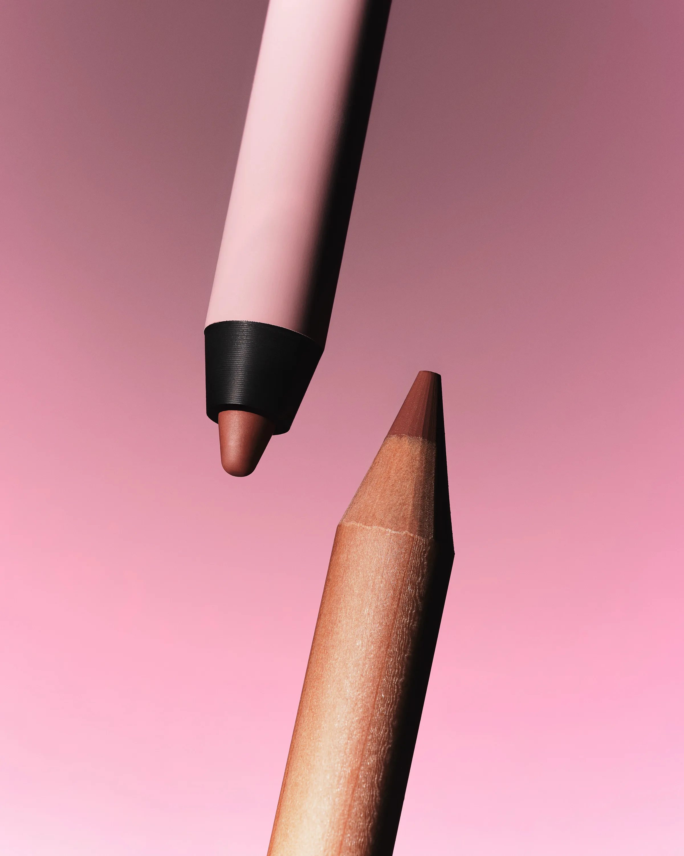 Two lip pencils, one pale pink and one wooden, tips facing each other against a pink gradient, for Kylie Cosmetics.