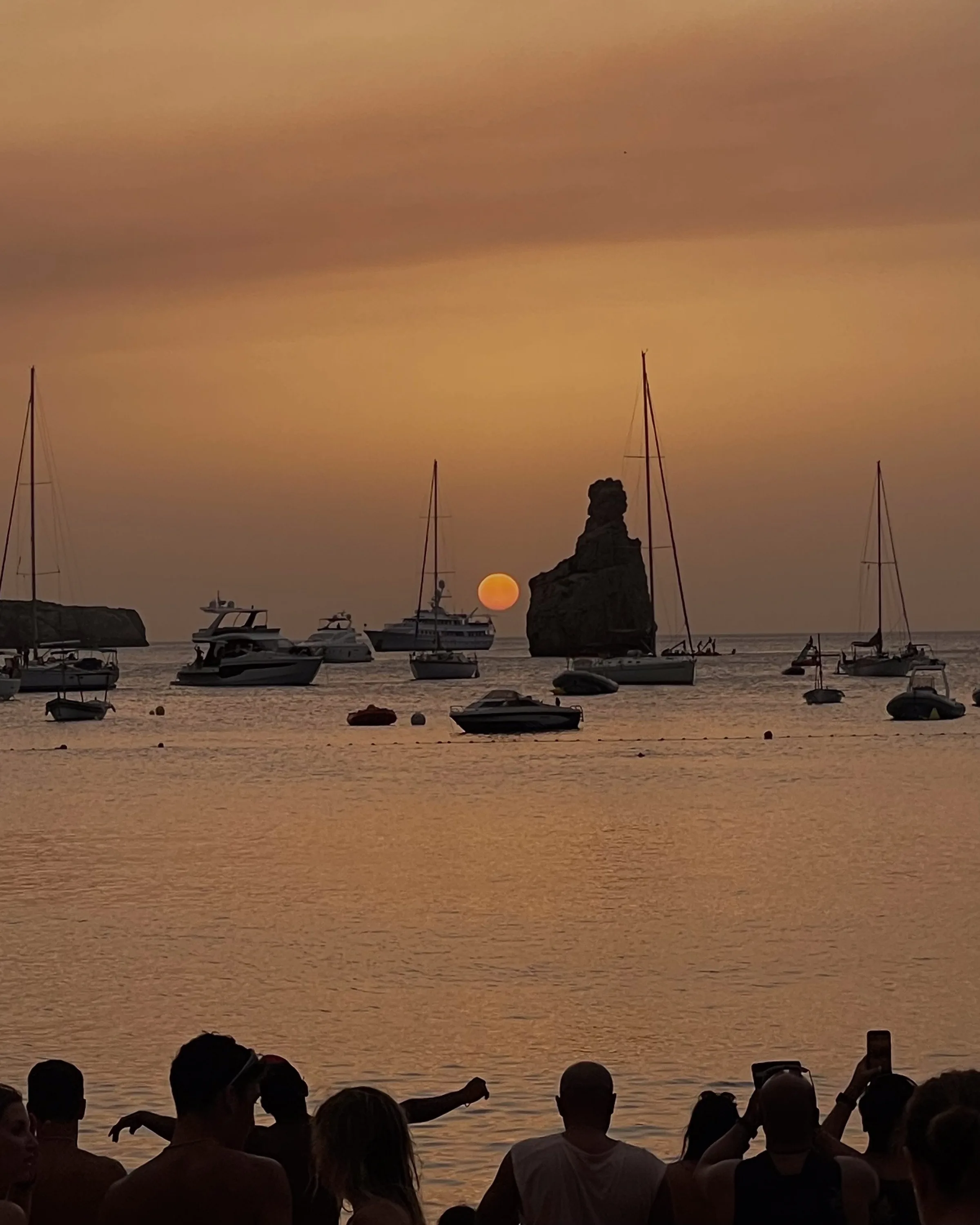 Crowd silhouetted on a beach watching an orange sunset over anchored sailboats and a rock spire in a hazy bay.