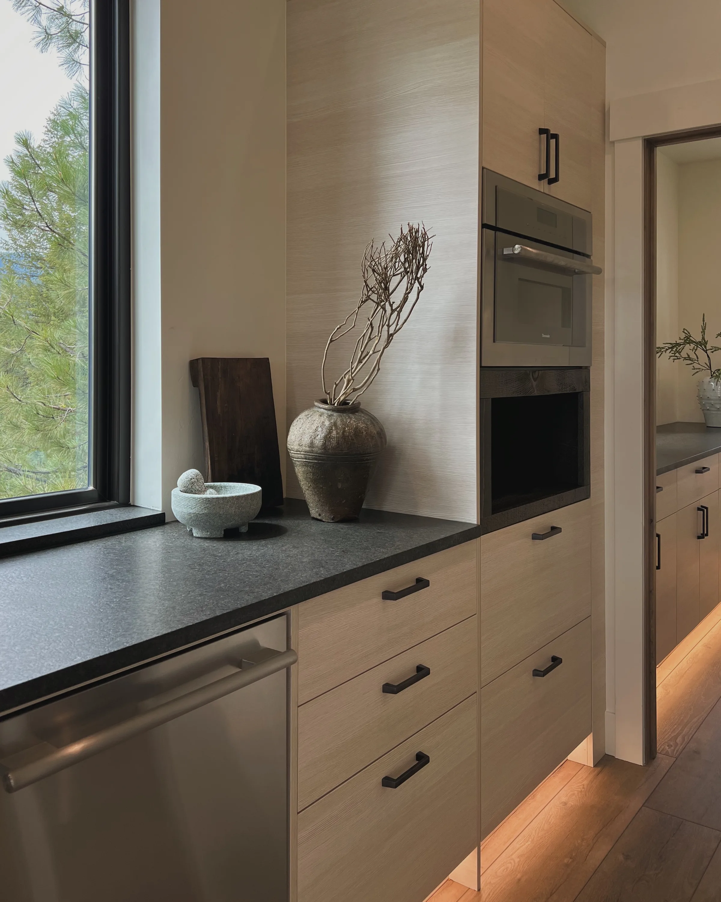 Modern kitchen with light wood cabinets, dark stone counter, stone vase of dry branches and mortar by a window.