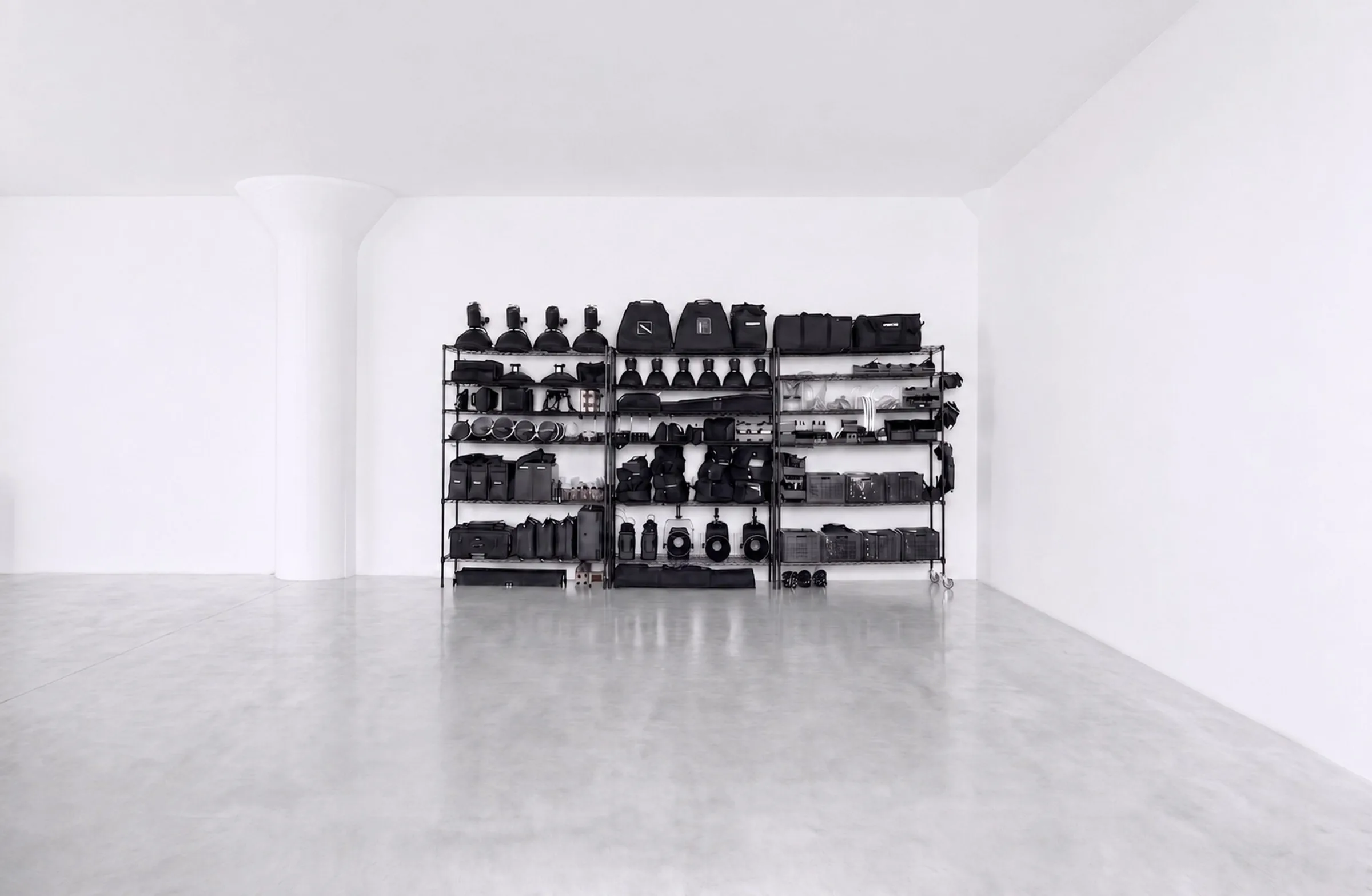 Black shelving stacked with lighting and grip gear against a white wall in Studio 1, Long Island City, with a concrete floor.