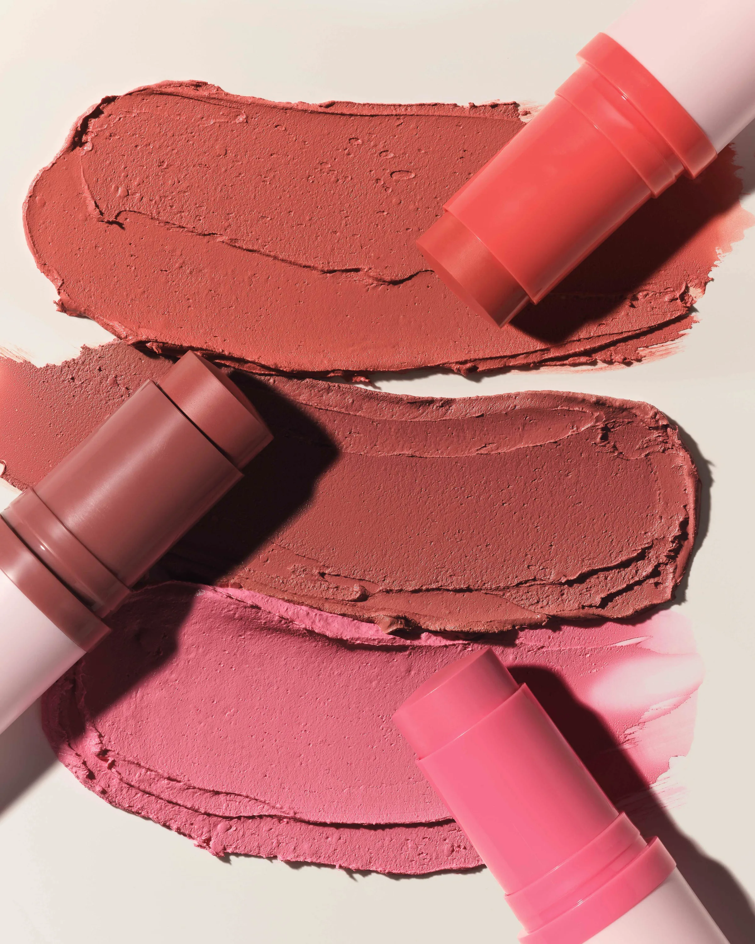 Three blush sticks in coral, mauve and pink beside creamy swatches on a pale background, for Kylie Cosmetics.