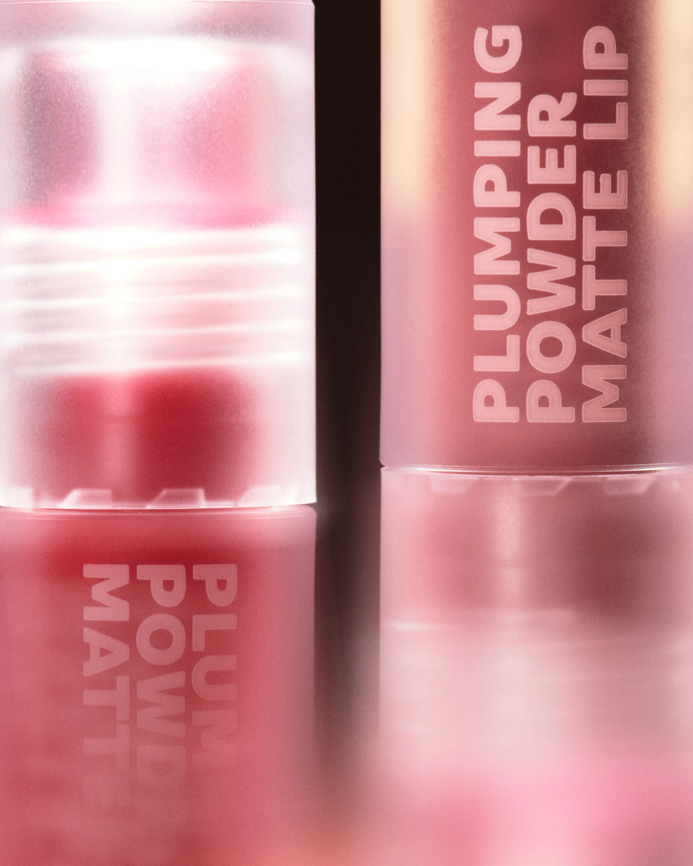 Close-up of two pink Plumping Powder Matte Lip tubes with frosted caps reflected on a glossy surface, for Kylie Cosmetics.