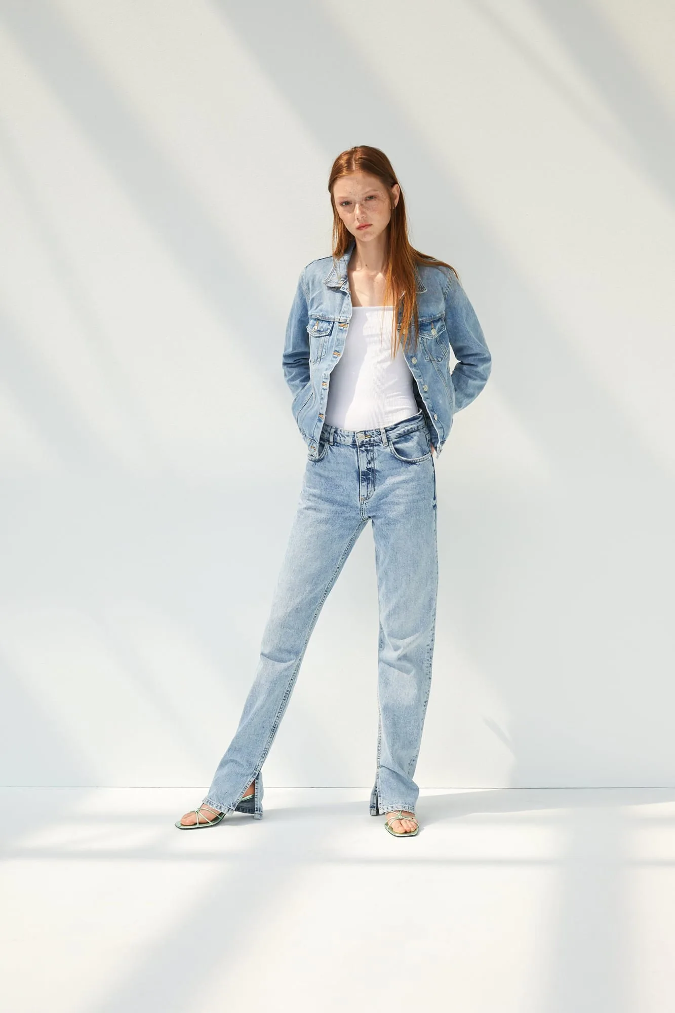 Red-haired model in a light wash denim jacket, white tank and split-hem jeans against a sunlit white wall, for Zara.