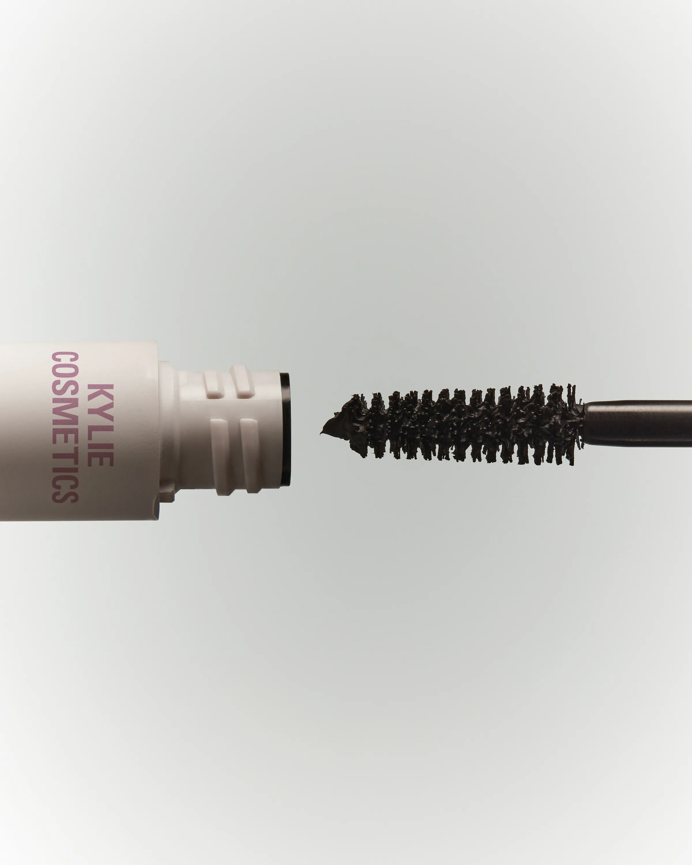 Still life of a mascara wand coated in black formula beside its open white tube on a pale grey backdrop, for Kylie Cosmetics.