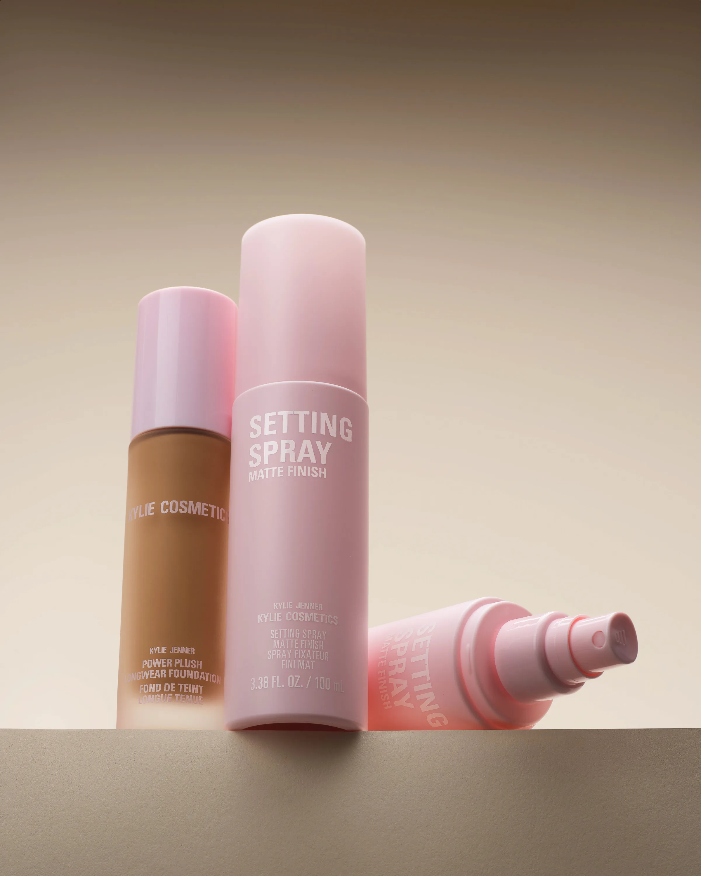 Pink matte setting spray bottles and Power Plush foundation on a beige ledge, one bottle lying on its side, for Kylie Cosmetics.