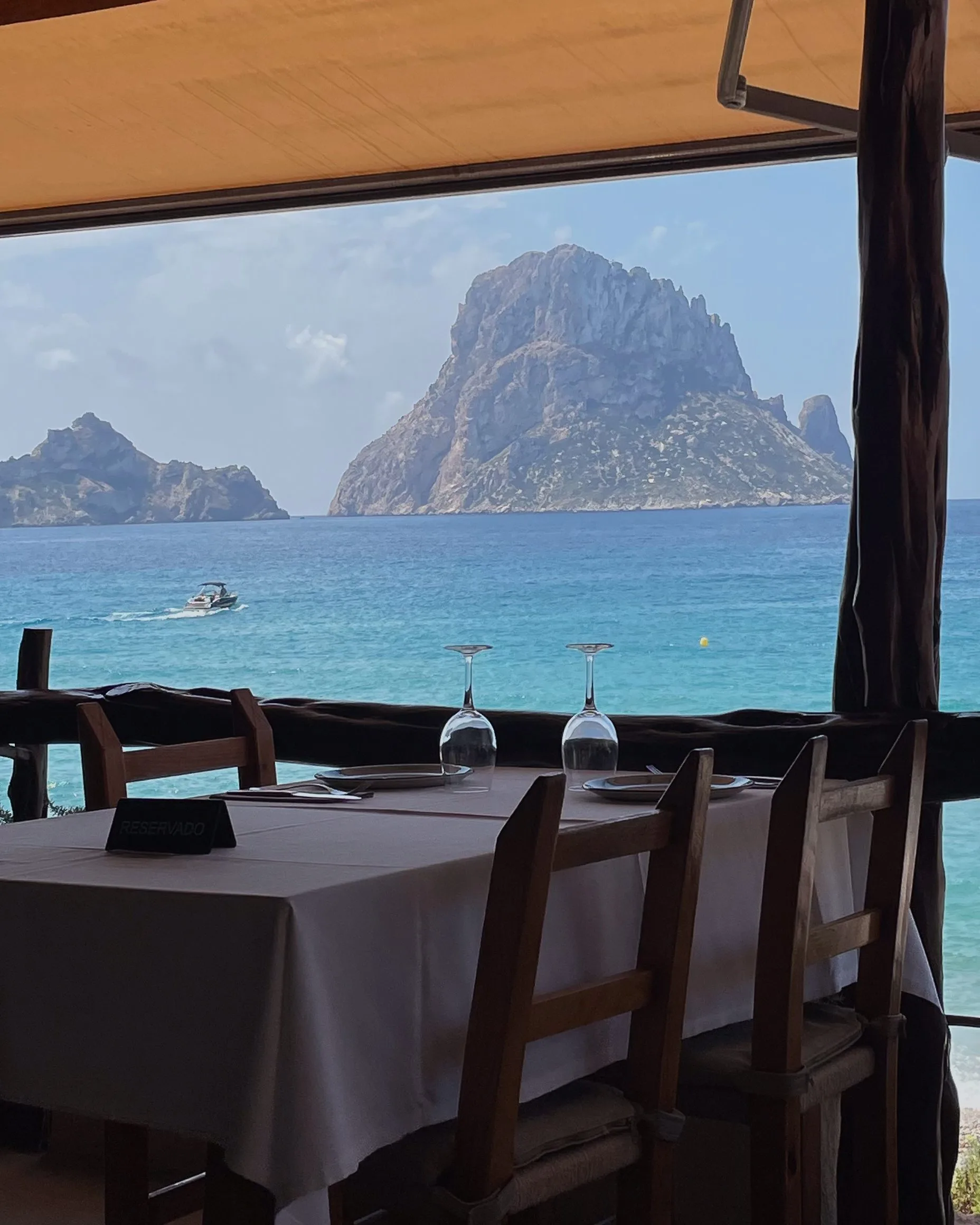 Restaurant table with wine glasses and a reserved sign overlooking turquoise sea and a rocky island, shot phone-style.