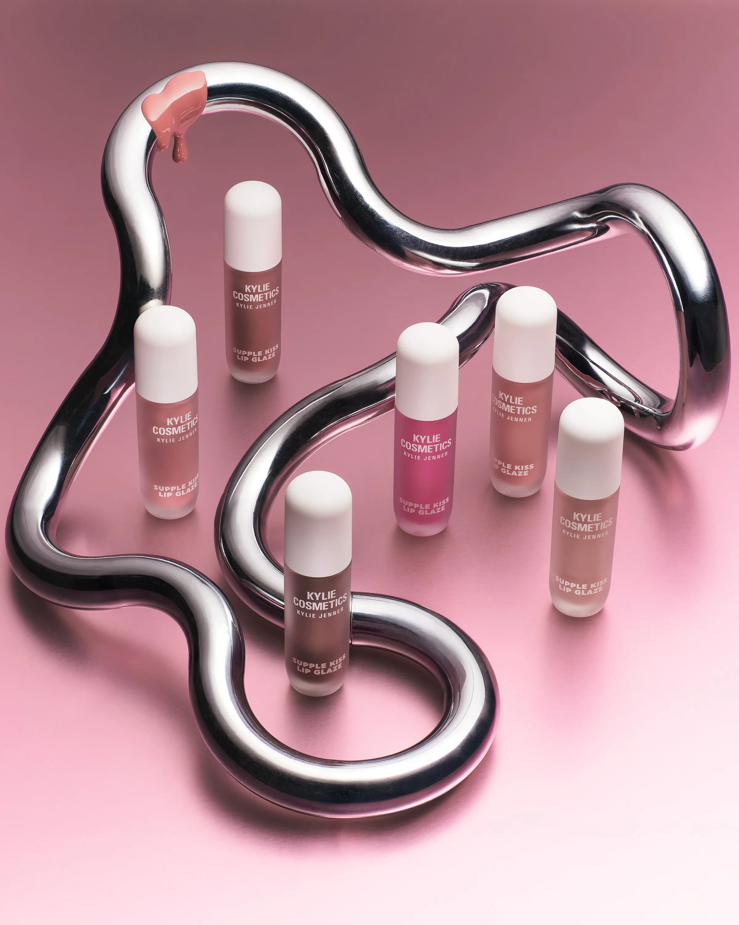 Supple Kiss Lip Glaze bottles in nude and pink shades inside a looping chrome tube on a pink surface, for Kylie Cosmetics.