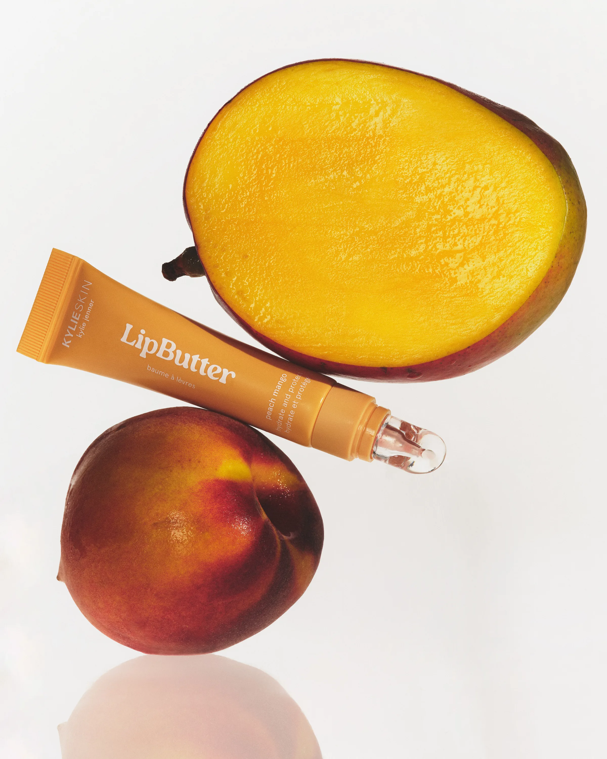 Still life of a peach mango Lip Butter tube balanced between a halved mango and a whole peach on white, for Kylie Cosmetics.
