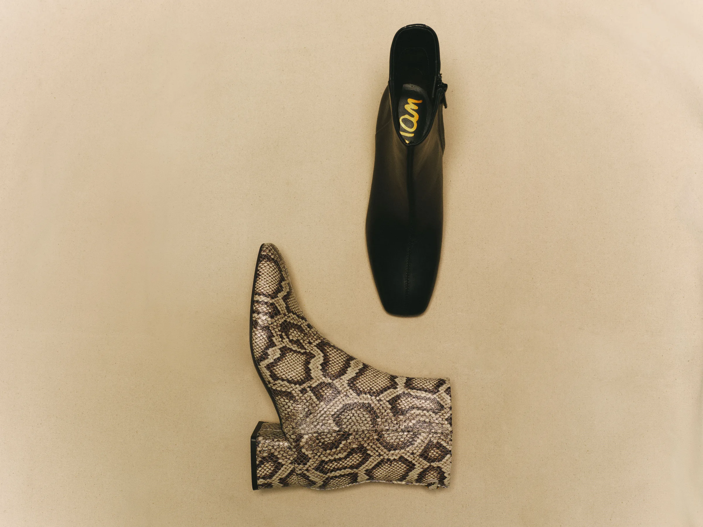 Black leather and snakeskin-print ankle boots laid flat on a beige backdrop, still life, for Sam Edelman.