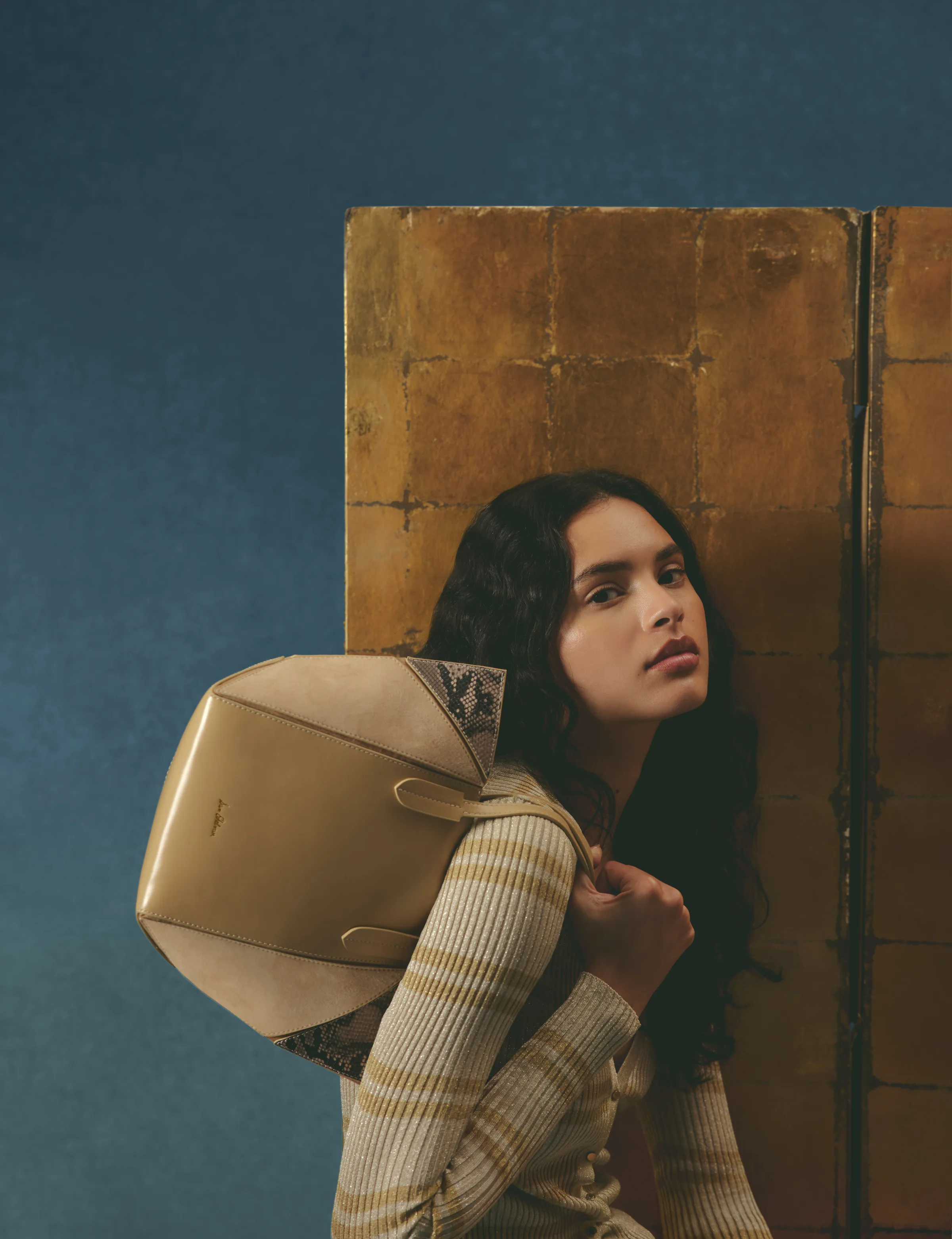 Model in a striped knit holds a beige leather and suede shoulder bag before a gold leaf screen, for Sam Edelman.