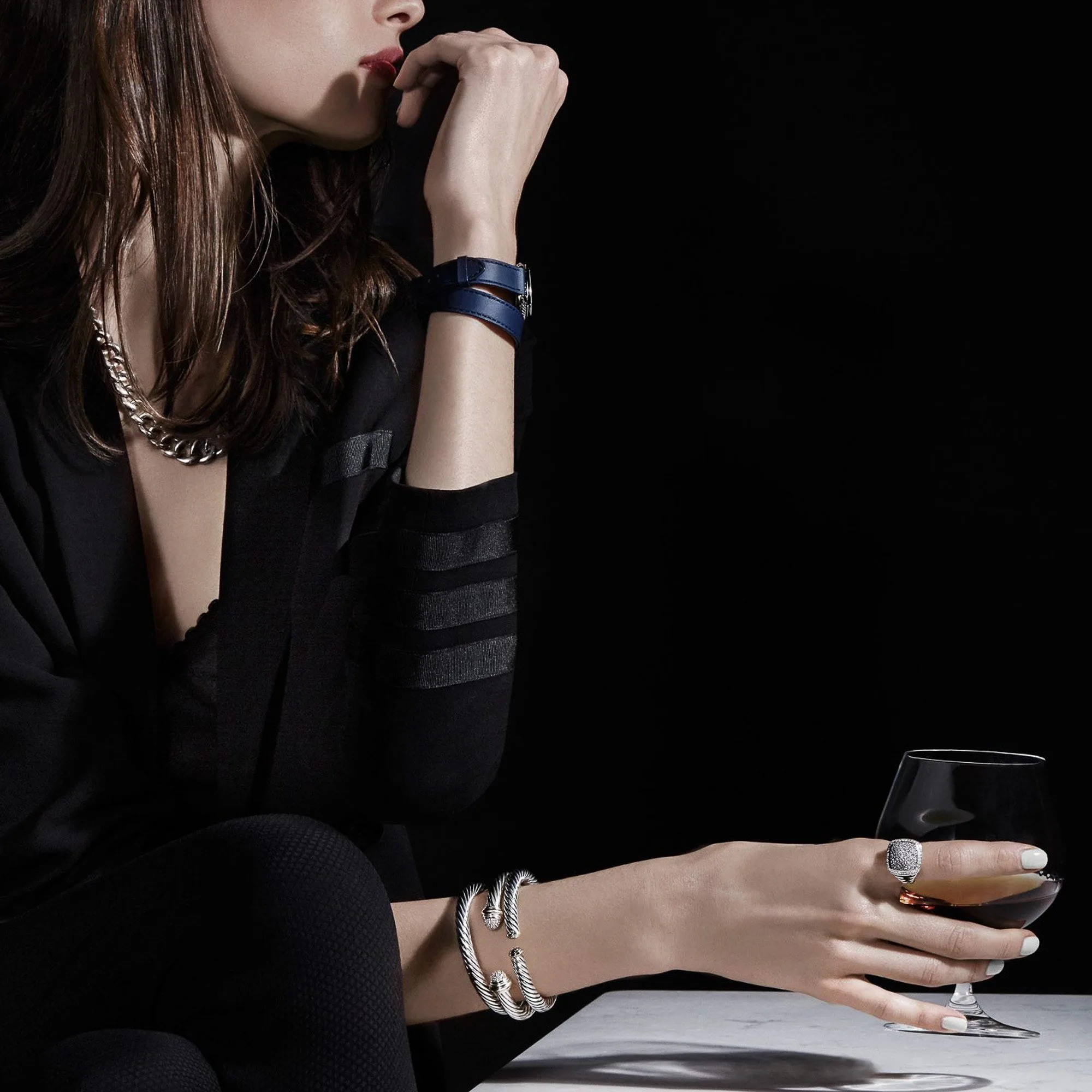 Model in black wearing silver cable bracelets, chain necklace and ring, holding a brandy glass on marble, for David Yurman.