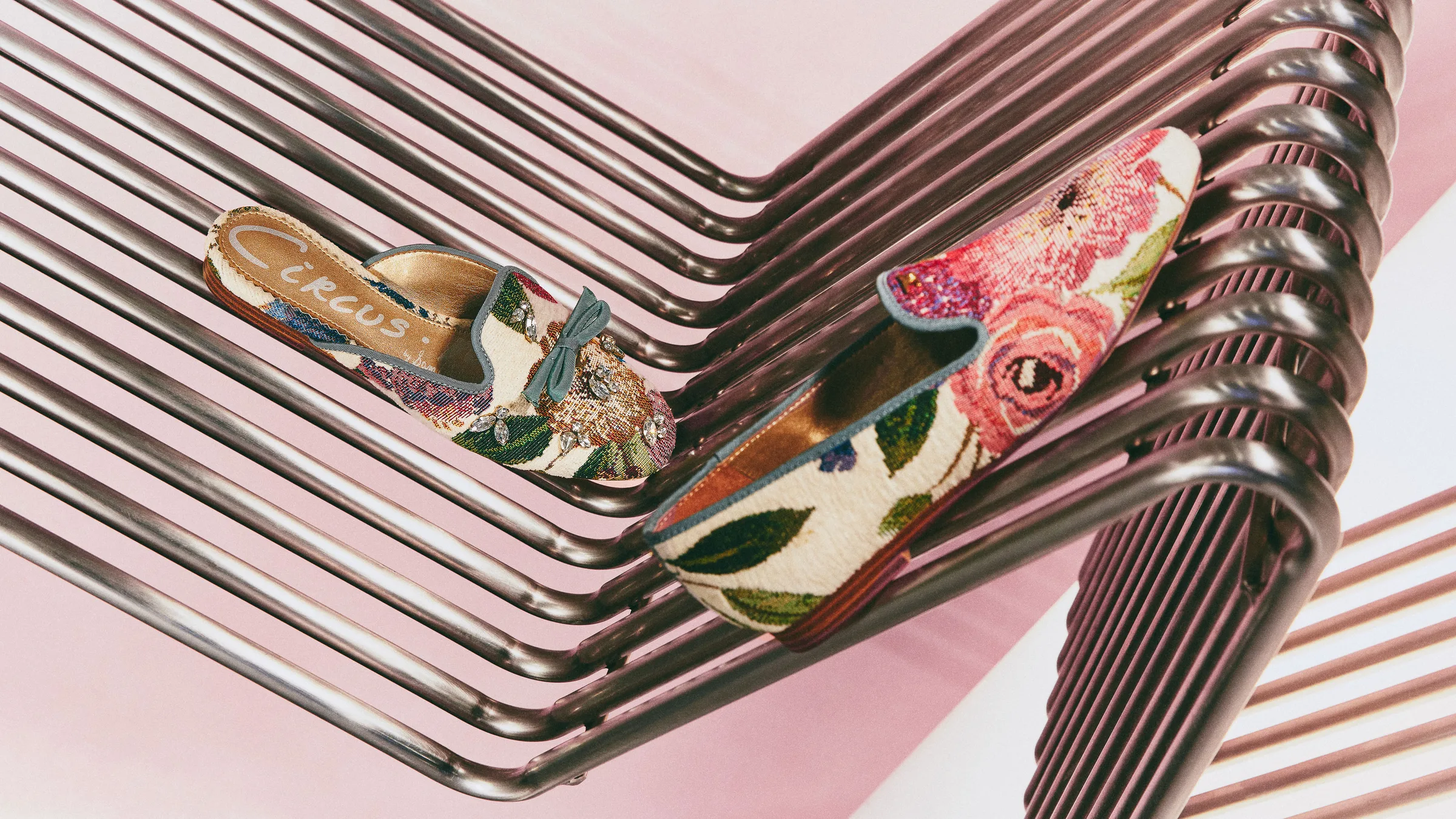 Floral tapestry flat mules, one with a jewelled bow, resting on a curved chrome wire rack against pink, for Circus NY.