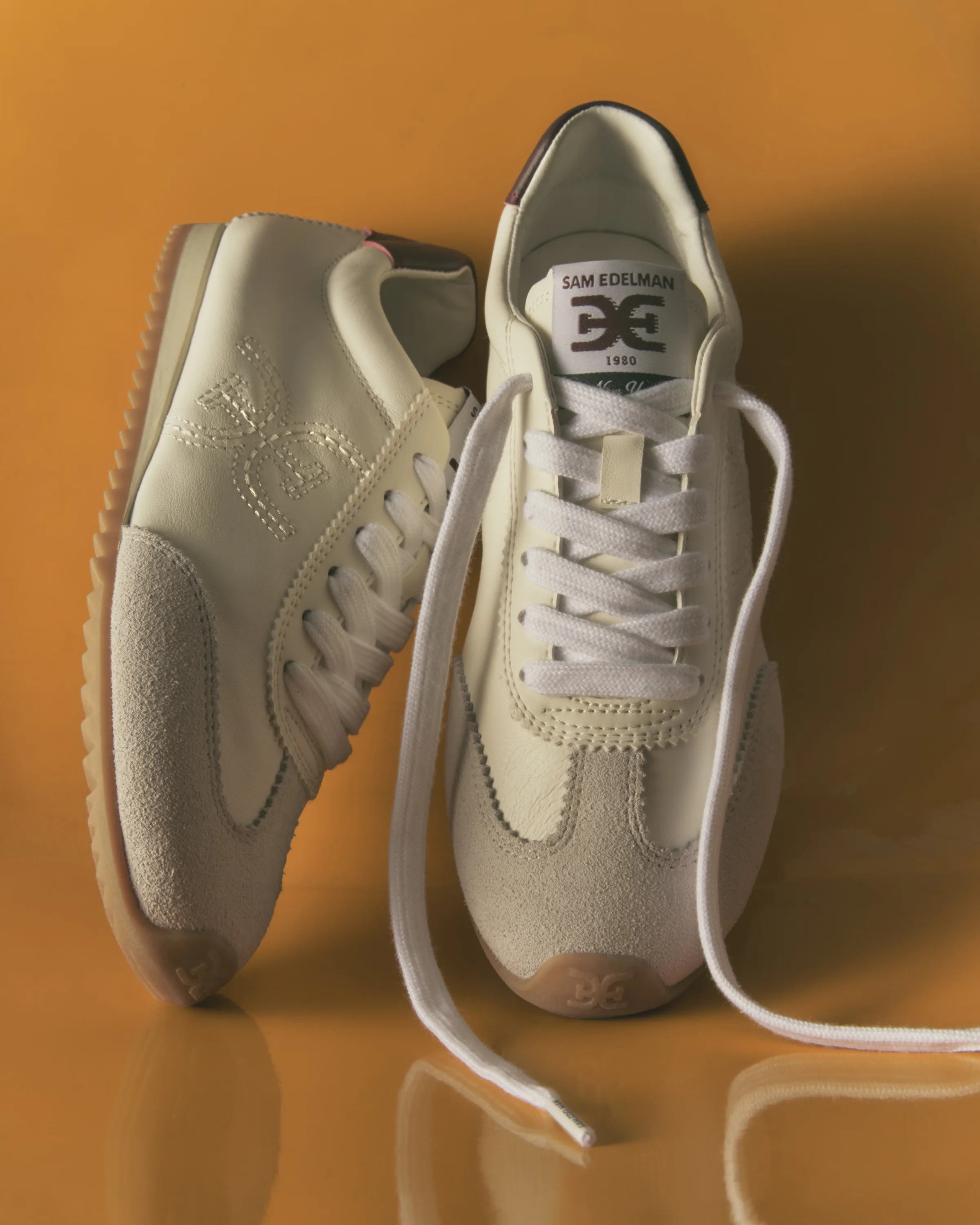 Pair of cream leather and suede retro sneakers with gum soles on a glossy orange surface, for Sam Edelman.