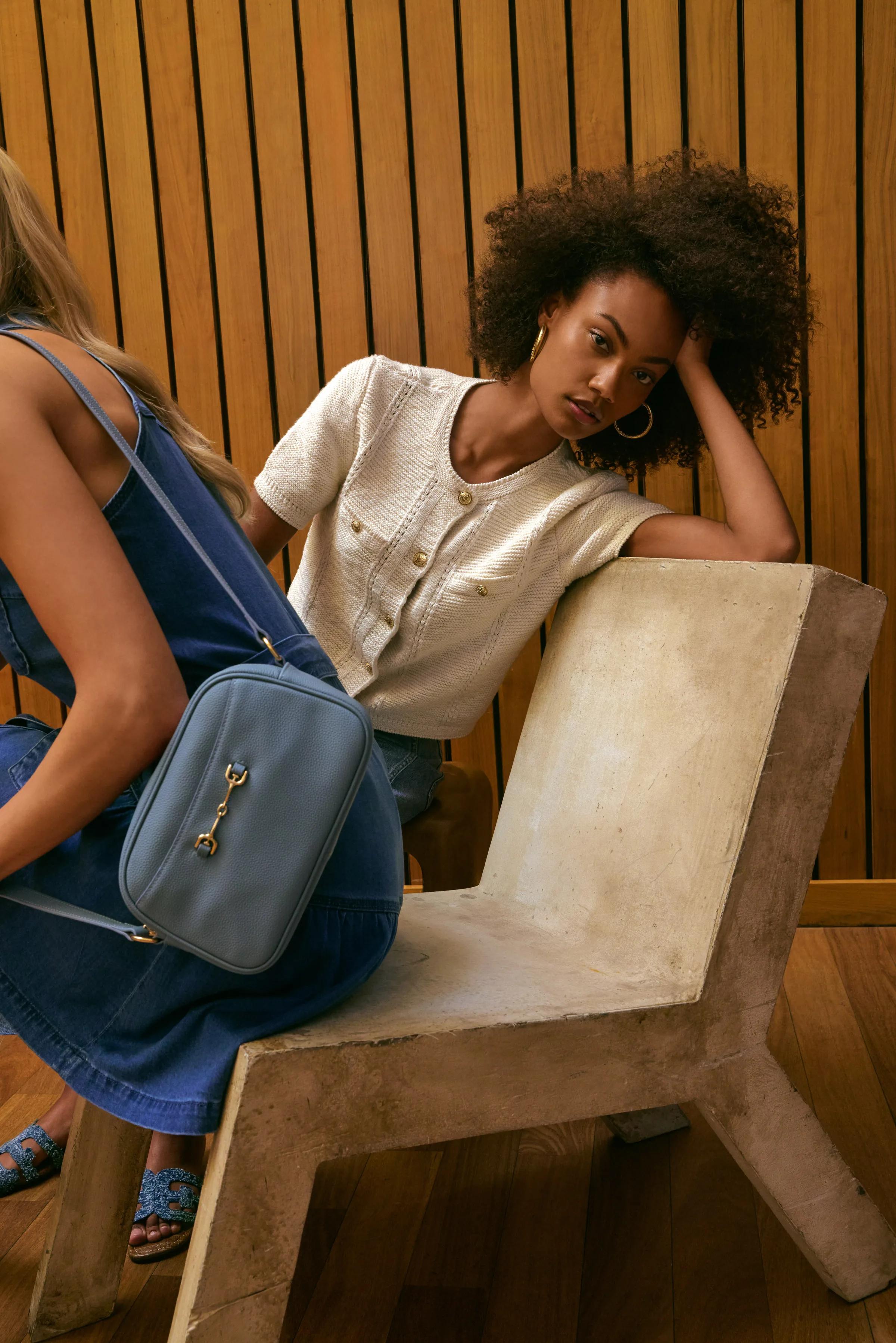 Two models on a concrete chair against wood slats, one in denim with a blue crossbody bag, for Sam Edelman.