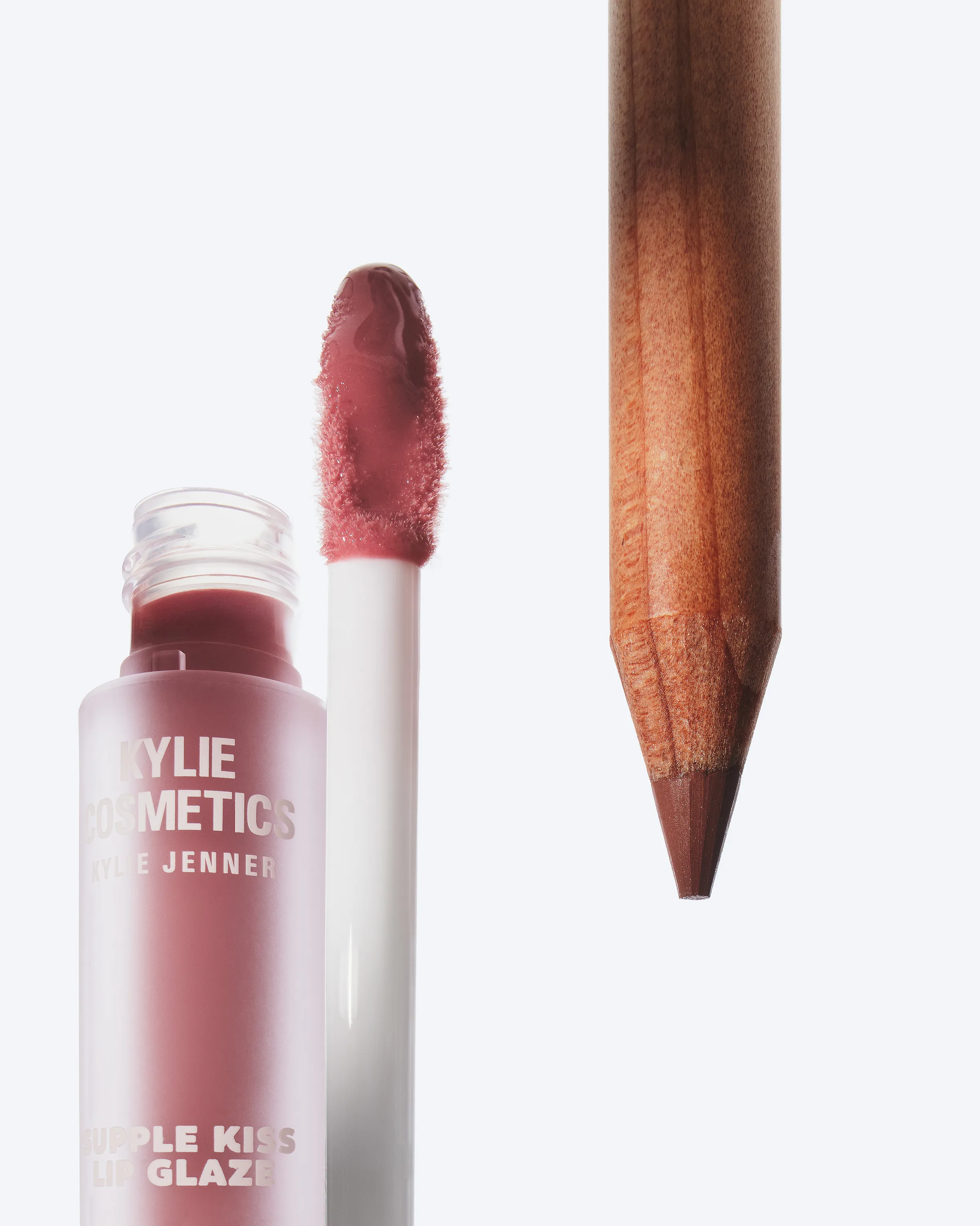 Still life of a mauve Supple Kiss lip glaze with its wand beside a wooden lip liner pencil on white, for Kylie Cosmetics.