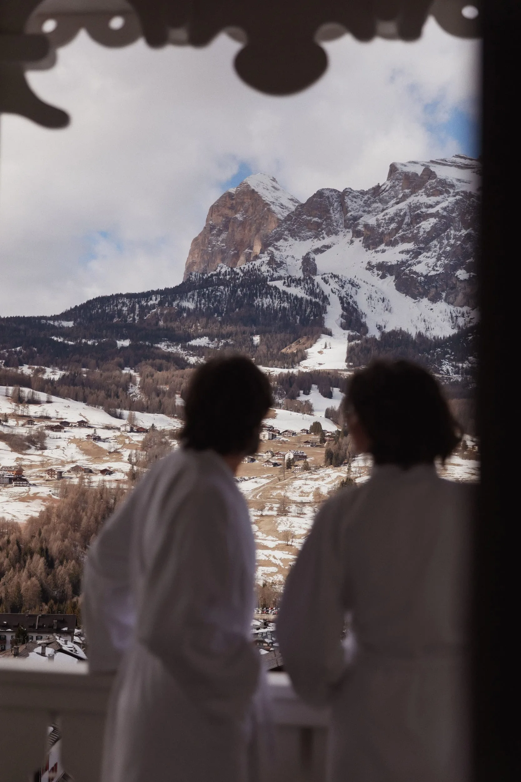 Two guests in white robes on a balcony looking out at snowy Dolomite peaks and a village, for Marriott.