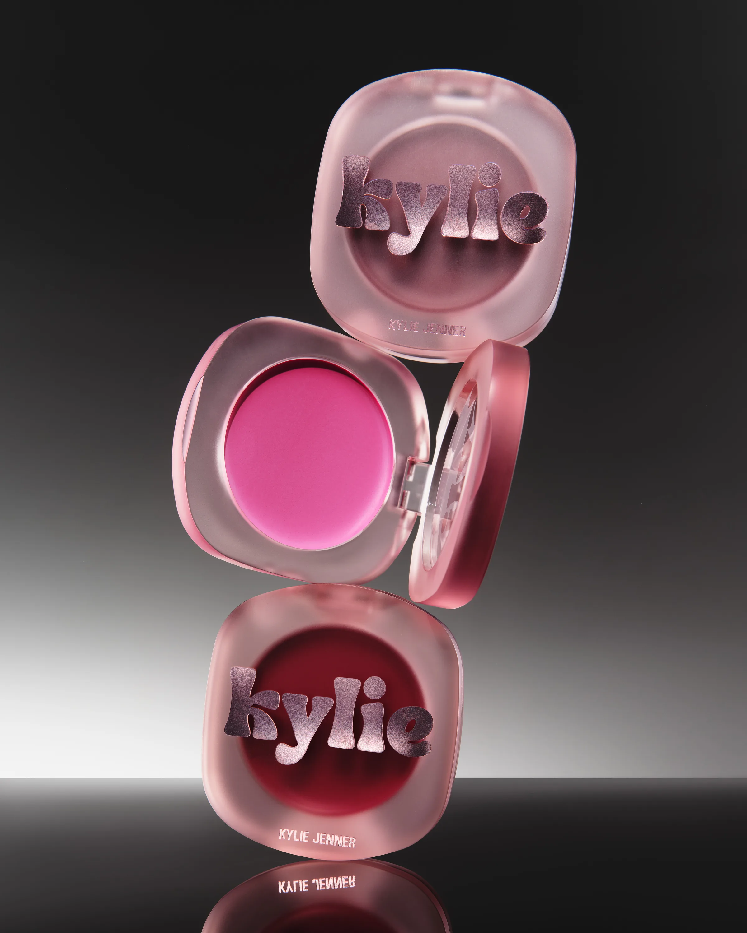 Three frosted pink glow balm compacts stacked mid-air, one open showing pink balm, on a black reflective surface, for Kylie Cosmetics.