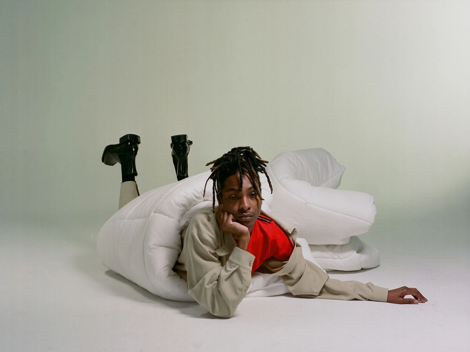 Model with locs in beige jacket and black boots lying on the floor wrapped in a white duvet, for VMAN.