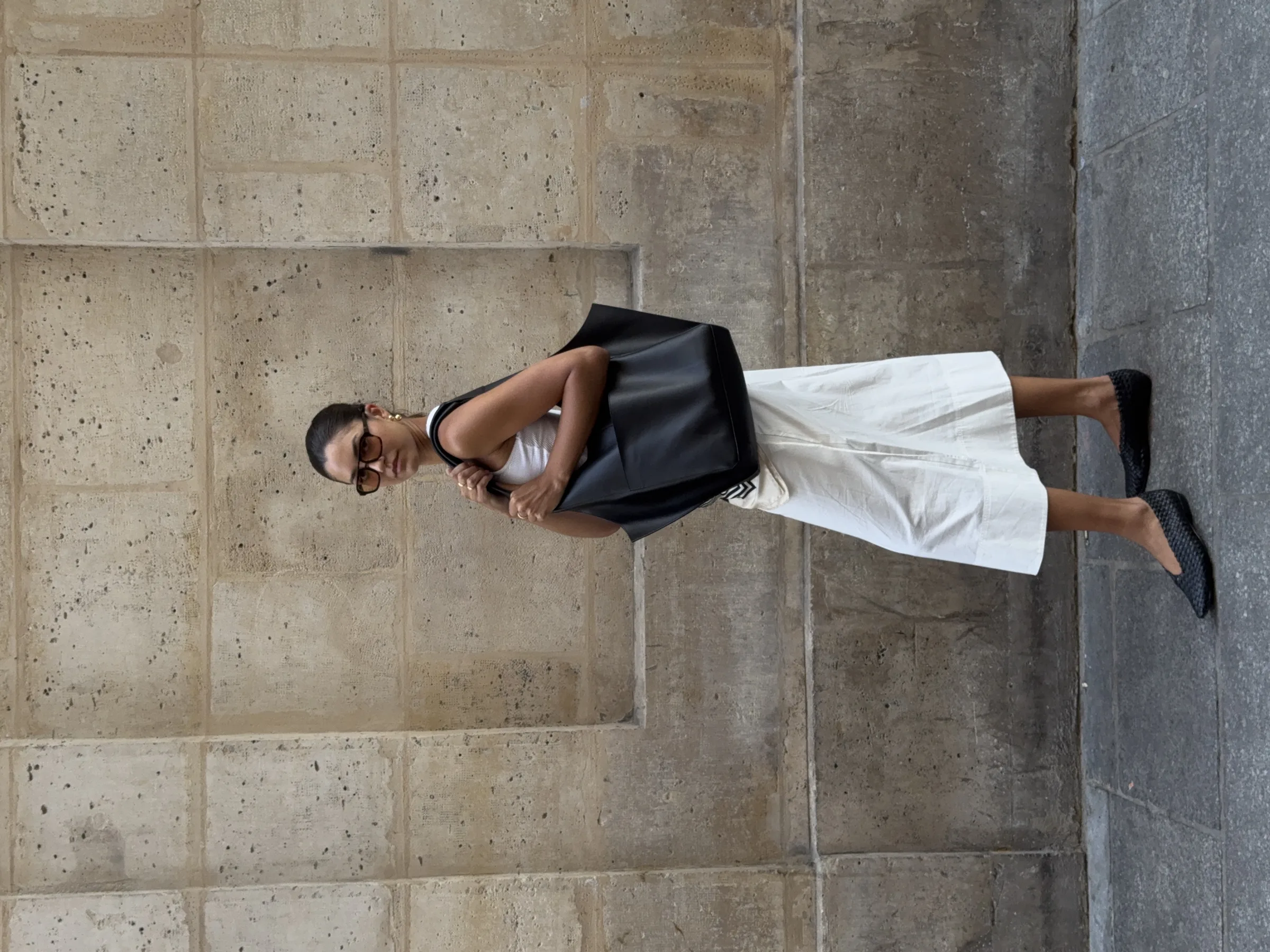 Woman in sunglasses, white tank and white midi skirt carrying a black leather tote against a limestone wall.