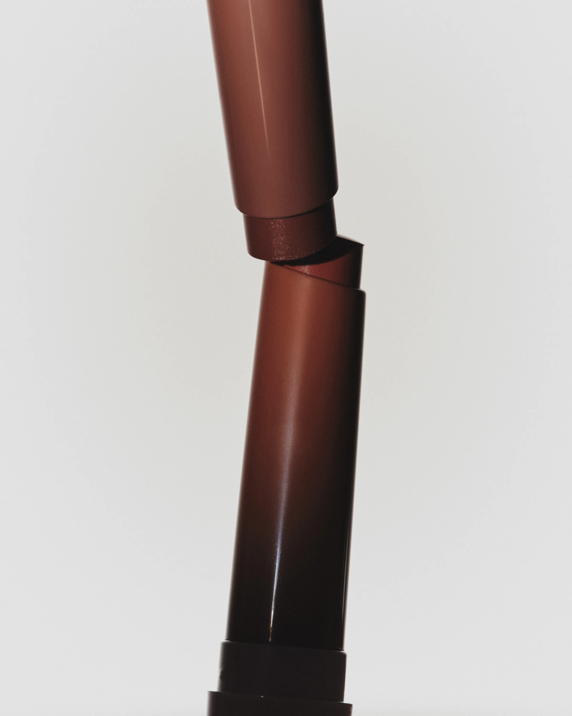 Two mocha-brown lip sticks stacked at an angle against a pale cream background, still life, for Kylie Cosmetics.