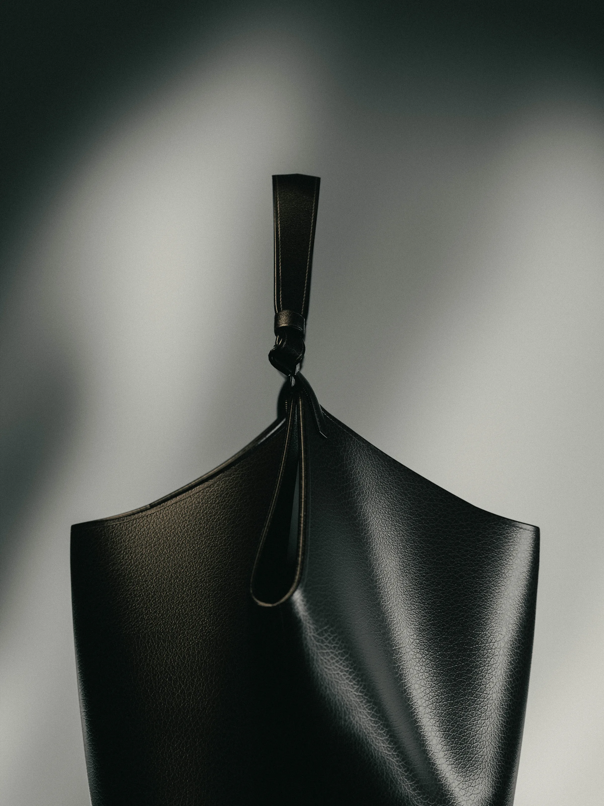CGI render of a black pebbled leather bucket bag with knotted strap against a shadowed grey wall, for Khaite.