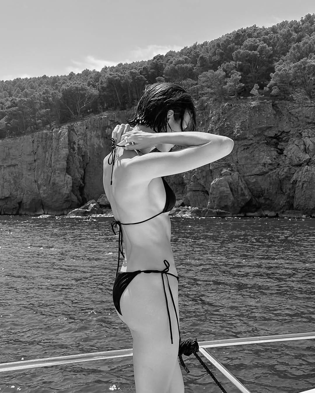 Black and white shot of a woman in a black string bikini tying her top on a boat deck beside rocky, wooded cliffs.