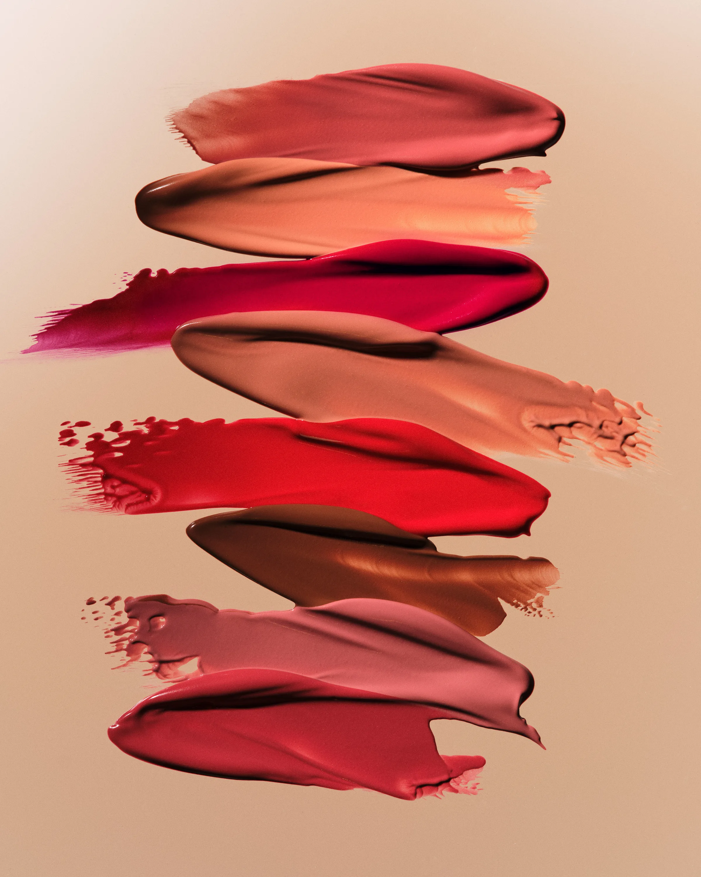 Eight lipstick swatches in reds, pinks and nudes smeared in a stack on a beige gradient background, for Kylie Cosmetics.