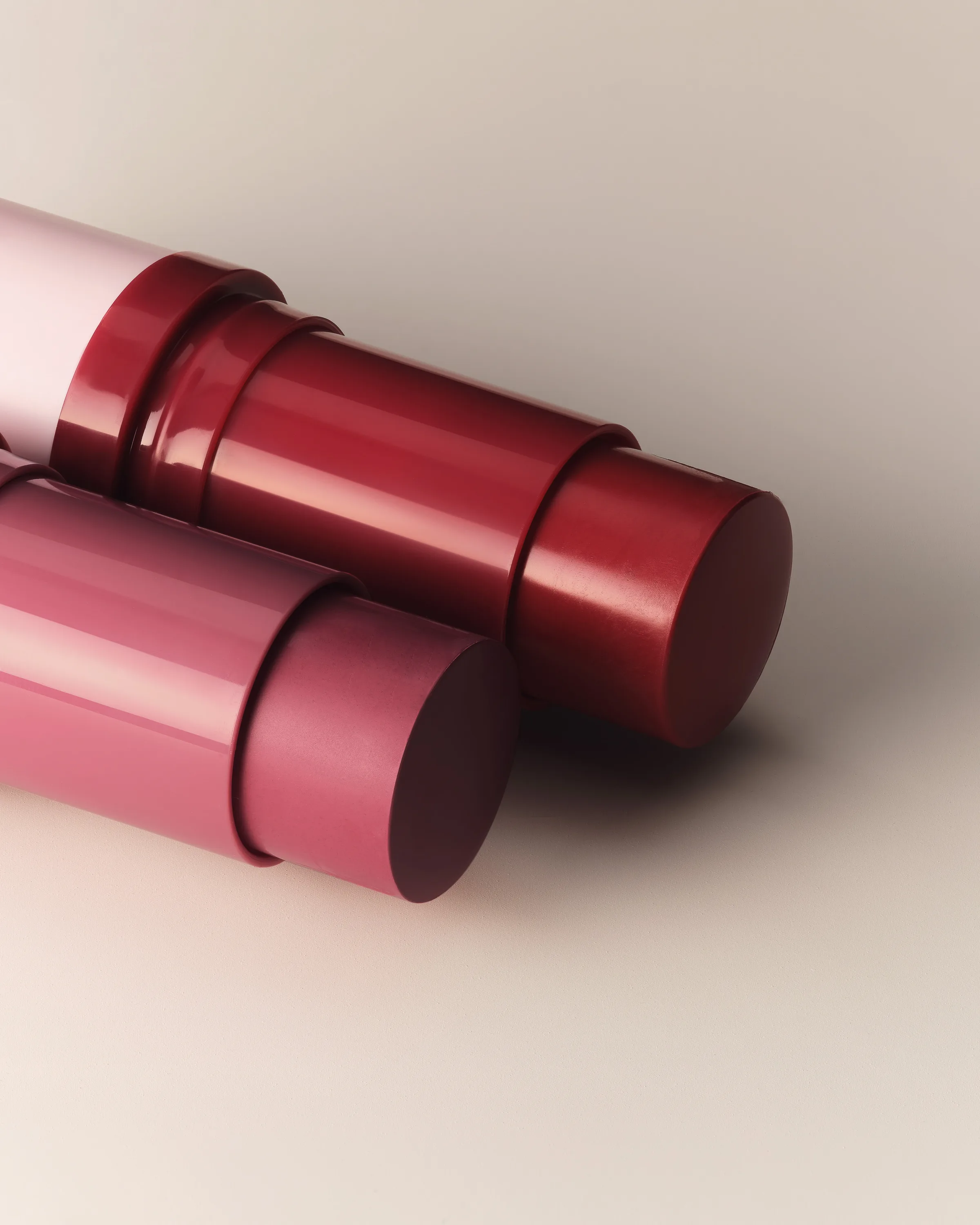 Still life close-up of two berry and pink twist-up lip sticks lying on a cream backdrop, for Kylie Cosmetics.