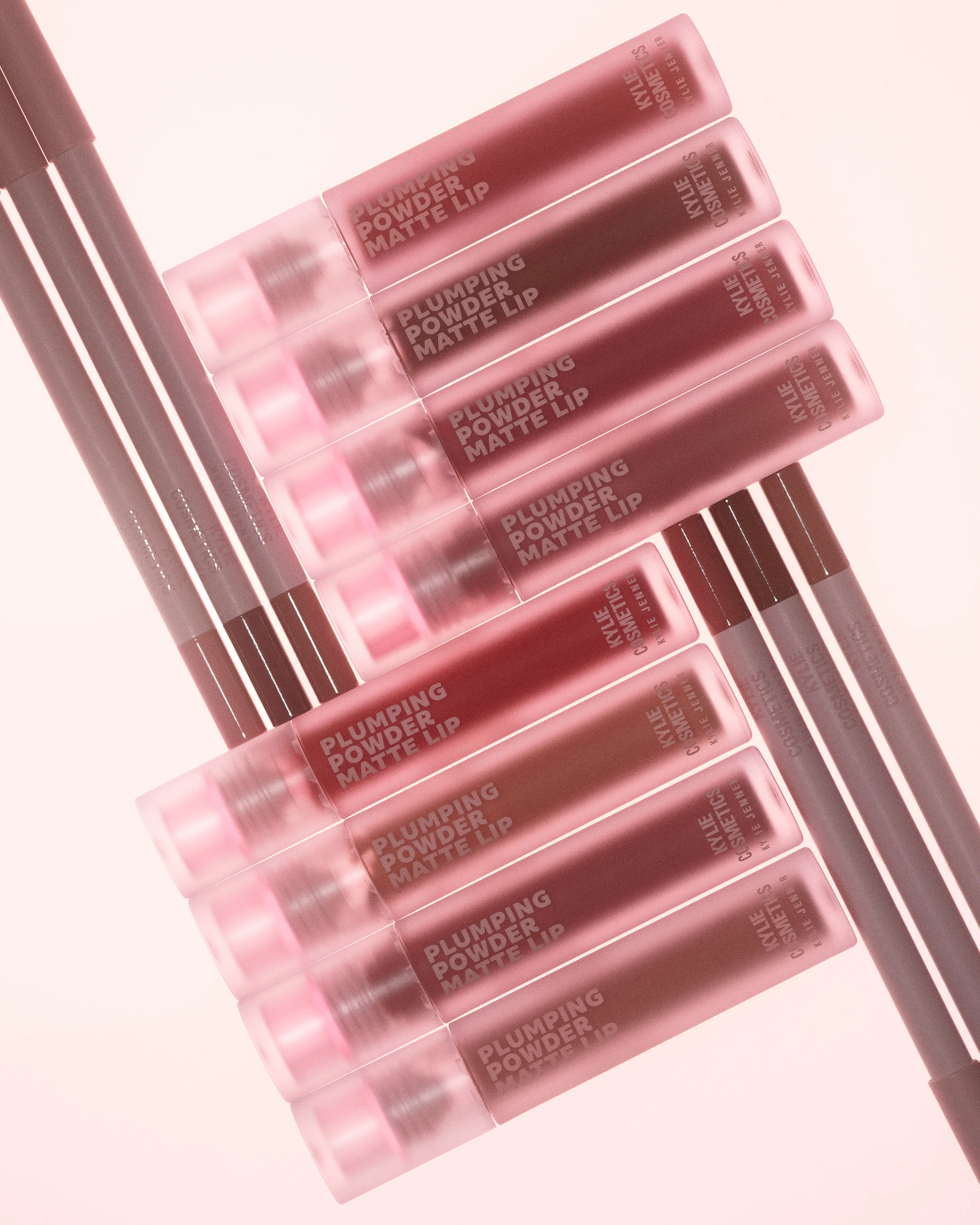 Plumping Powder Matte Lip tubes in rose and mauve shades fanned over lip liners on a pale pink background, for Kylie Cosmetics.