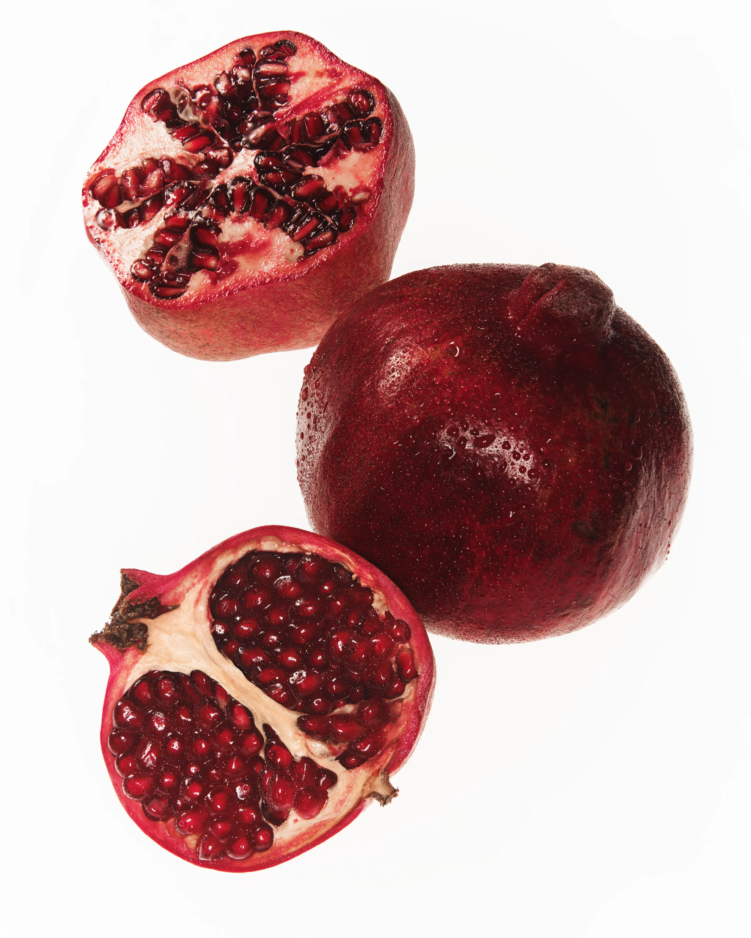 Still life of a whole pomegranate with dewdrops beside two cut halves showing red seeds on white, for Kylie Cosmetics.