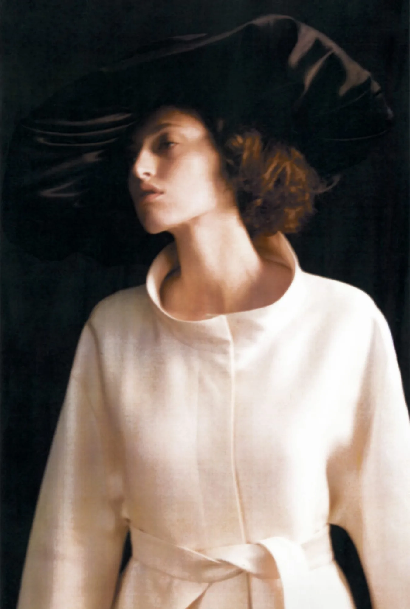 Model in a wide black satin hat and belted cream coat against a dark backdrop, shot by Amber Witcomb.
