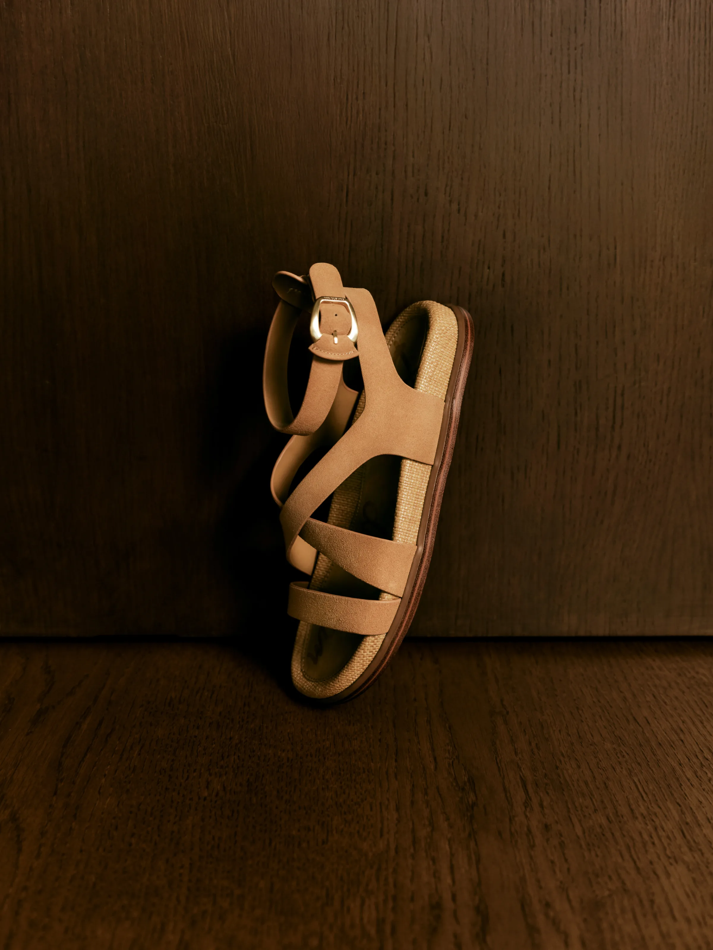 Tan suede strappy sandal with gold buckle and woven footbed, propped against dark wood panelling, for Sam Edelman.