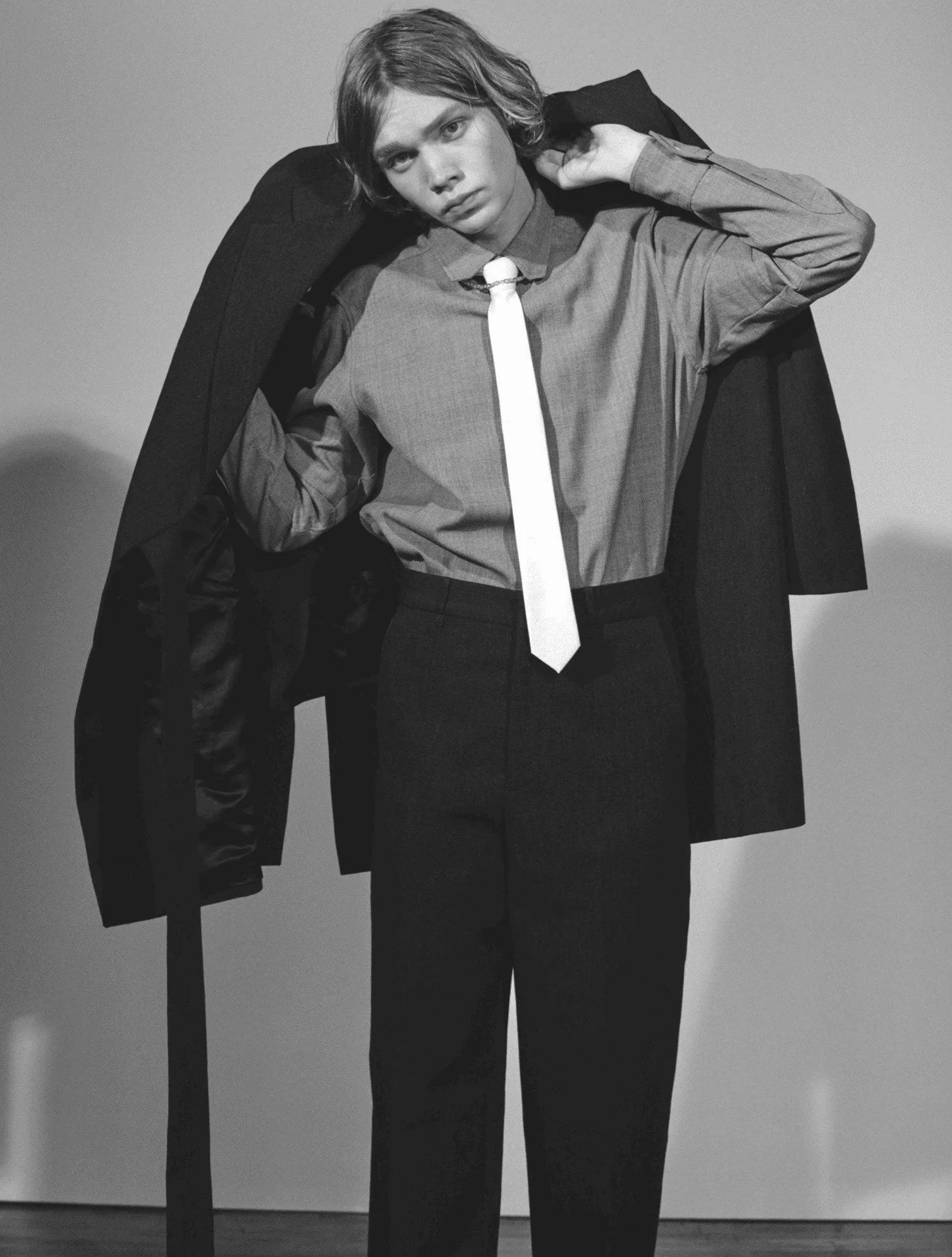 Black and white portrait of a model in a grey shirt, white tie and dark trousers, coat slung over shoulder, for L'Officiel Hommes.