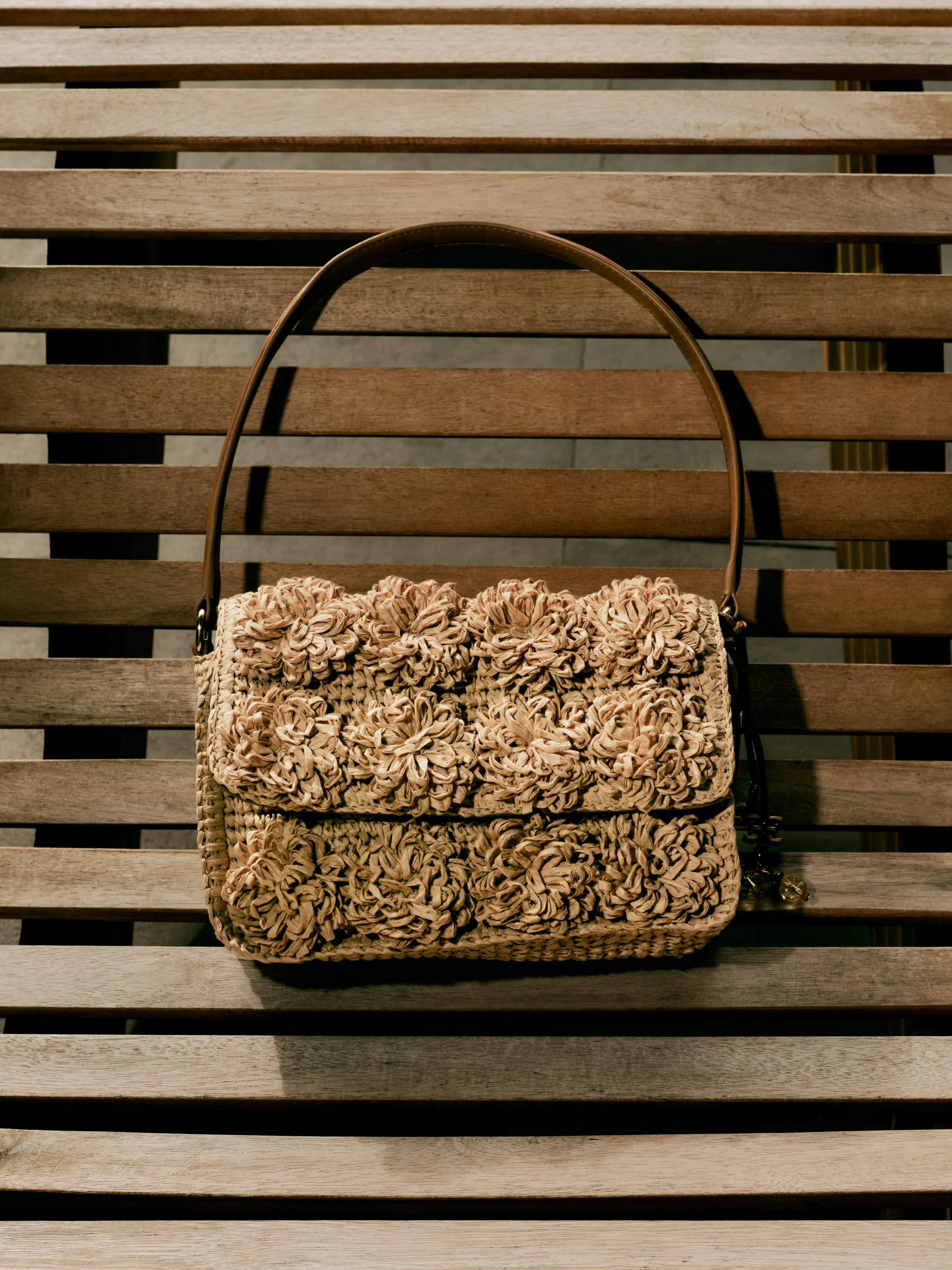 Woven raffia shoulder bag with flower appliqués and tan leather strap on a sunlit wooden slat bench, for Sam Edelman.