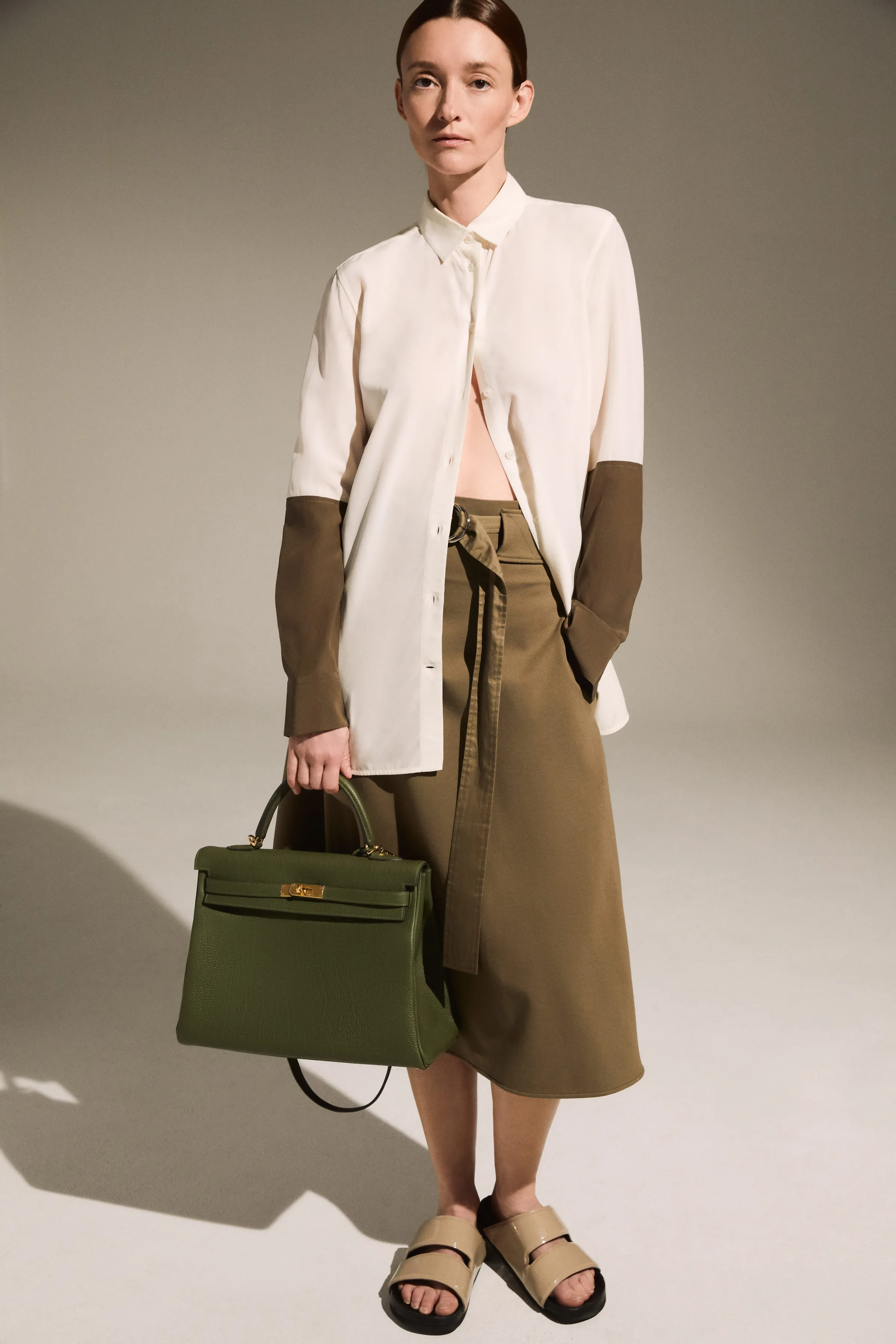 Model in cream shirt with olive cuffs and belted khaki skirt holding an olive leather handbag, for The RealReal.