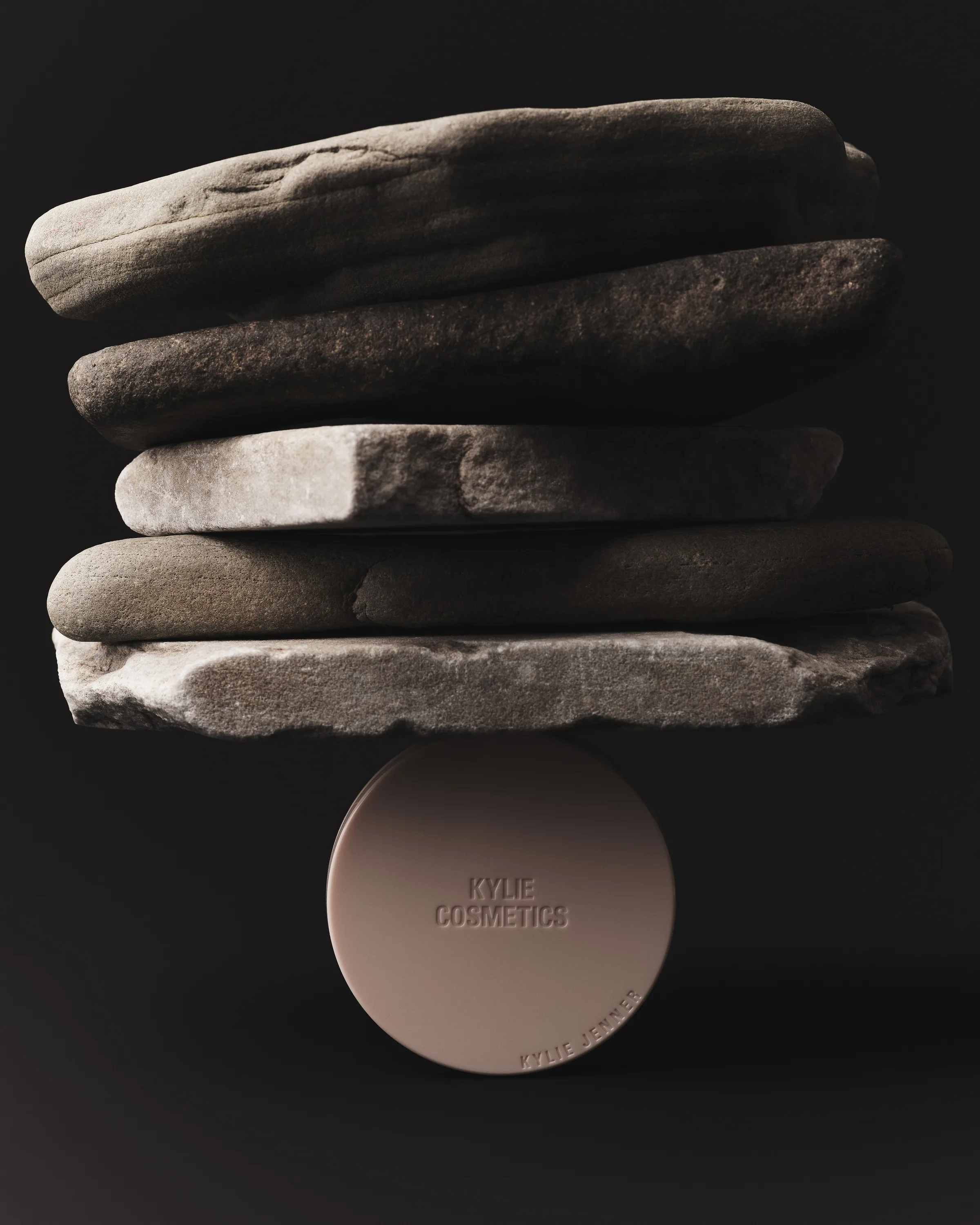 Taupe powder compact balancing a stack of flat stones against a dark background, for Kylie Cosmetics.