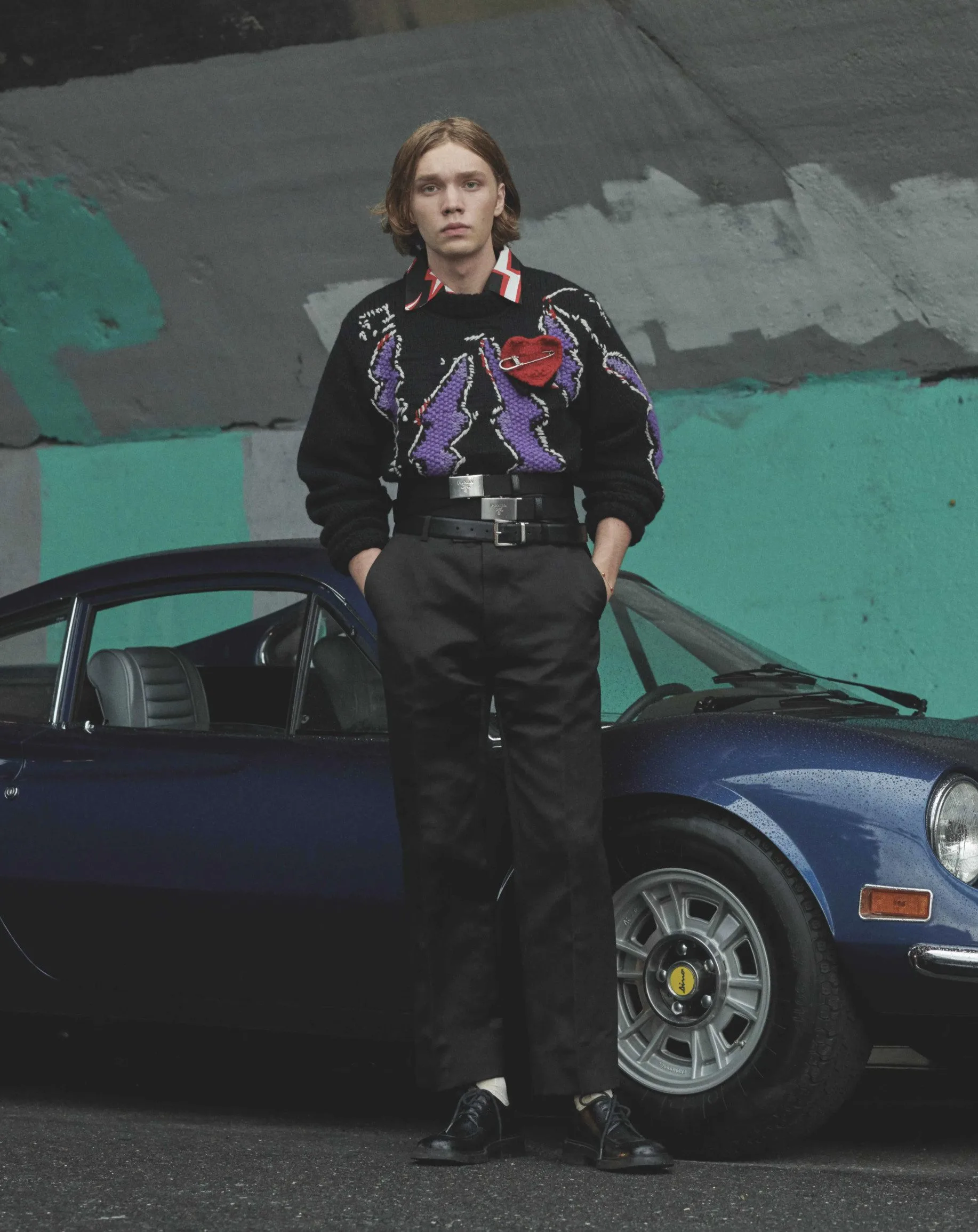 Model in purple-patterned black sweater and black trousers leaning on a blue vintage Ferrari, for L'Officiel Hommes.