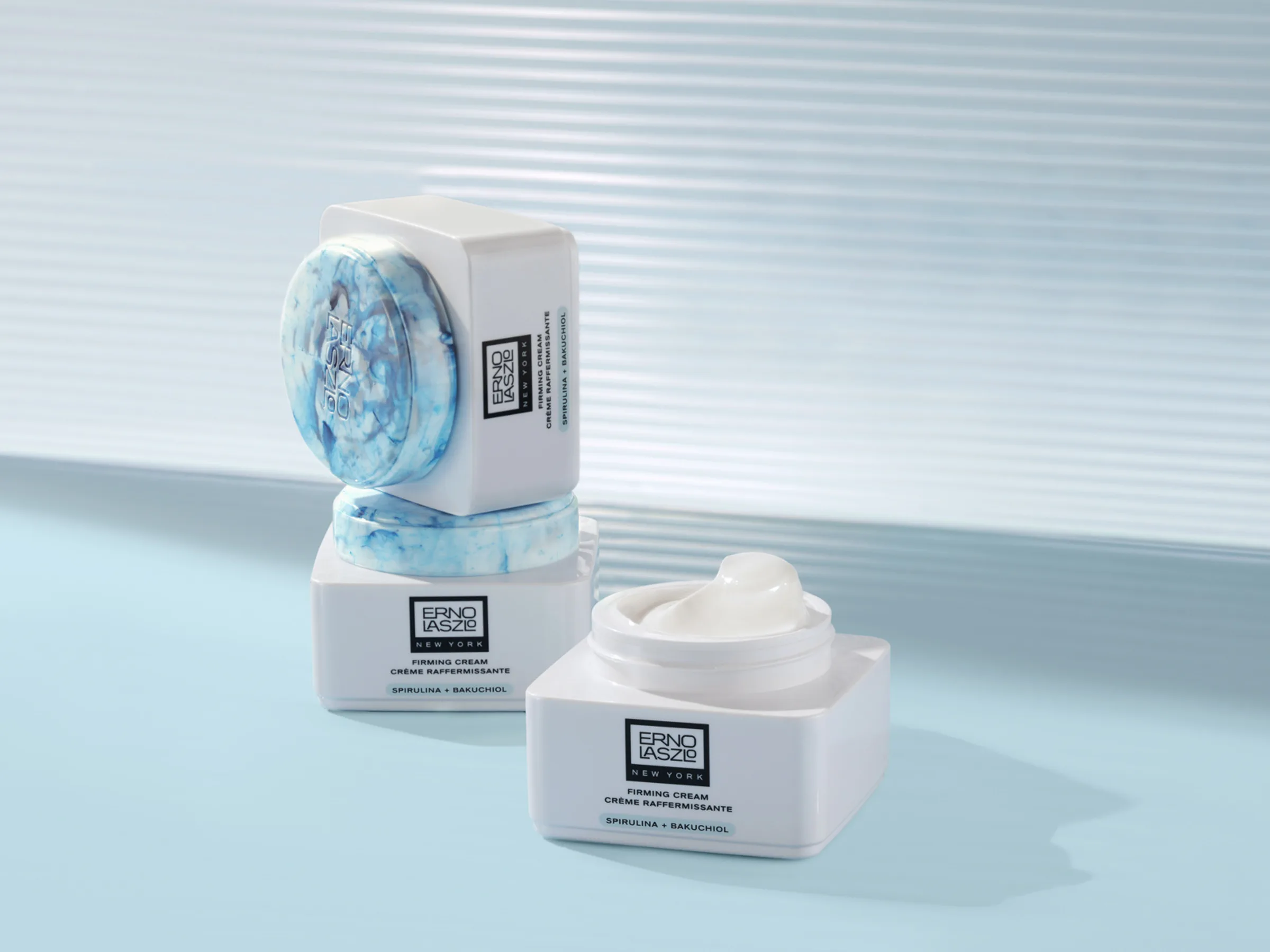 White Firming Cream jars with marbled blue lids, one open, stacked on a pale blue set with ribbed glass, for Erno Laszlo.