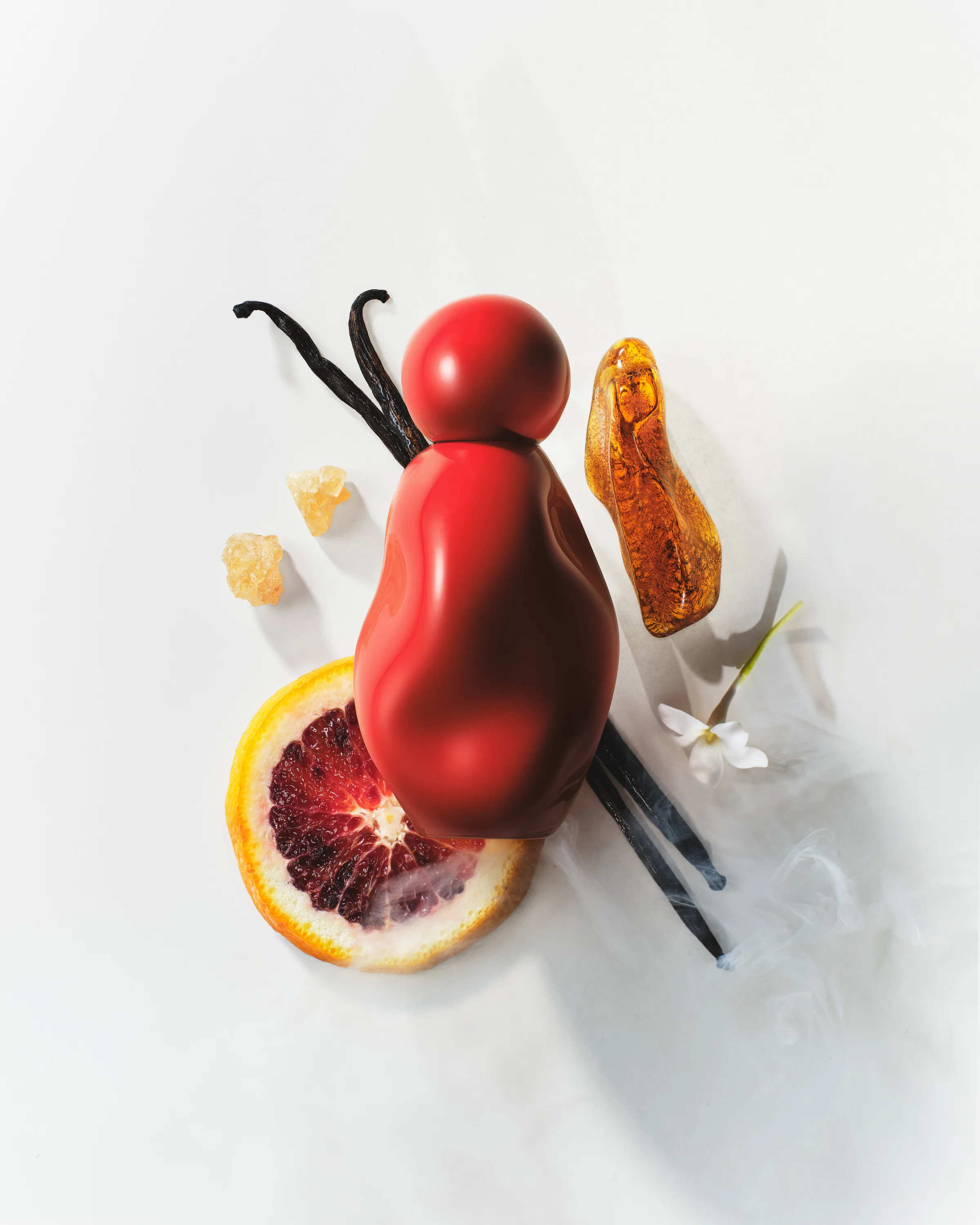 Red sculpted perfume bottle lying among vanilla pods, blood orange slice, amber resin and white flower, for Kylie Cosmetics.