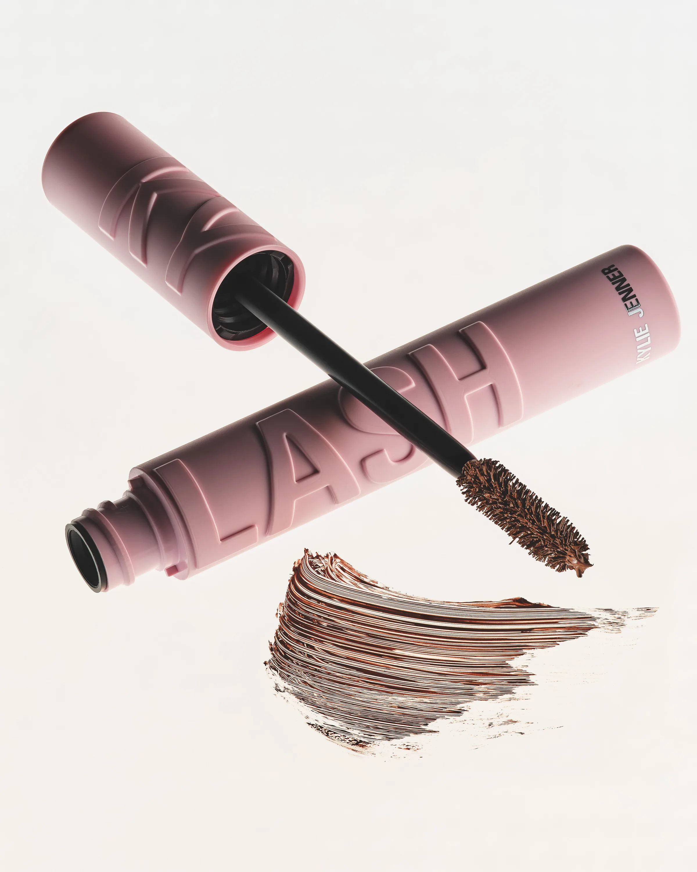 Pink Kylash mascara tube and wand floating over a brown mascara swatch on a white background, for Kylie Cosmetics.