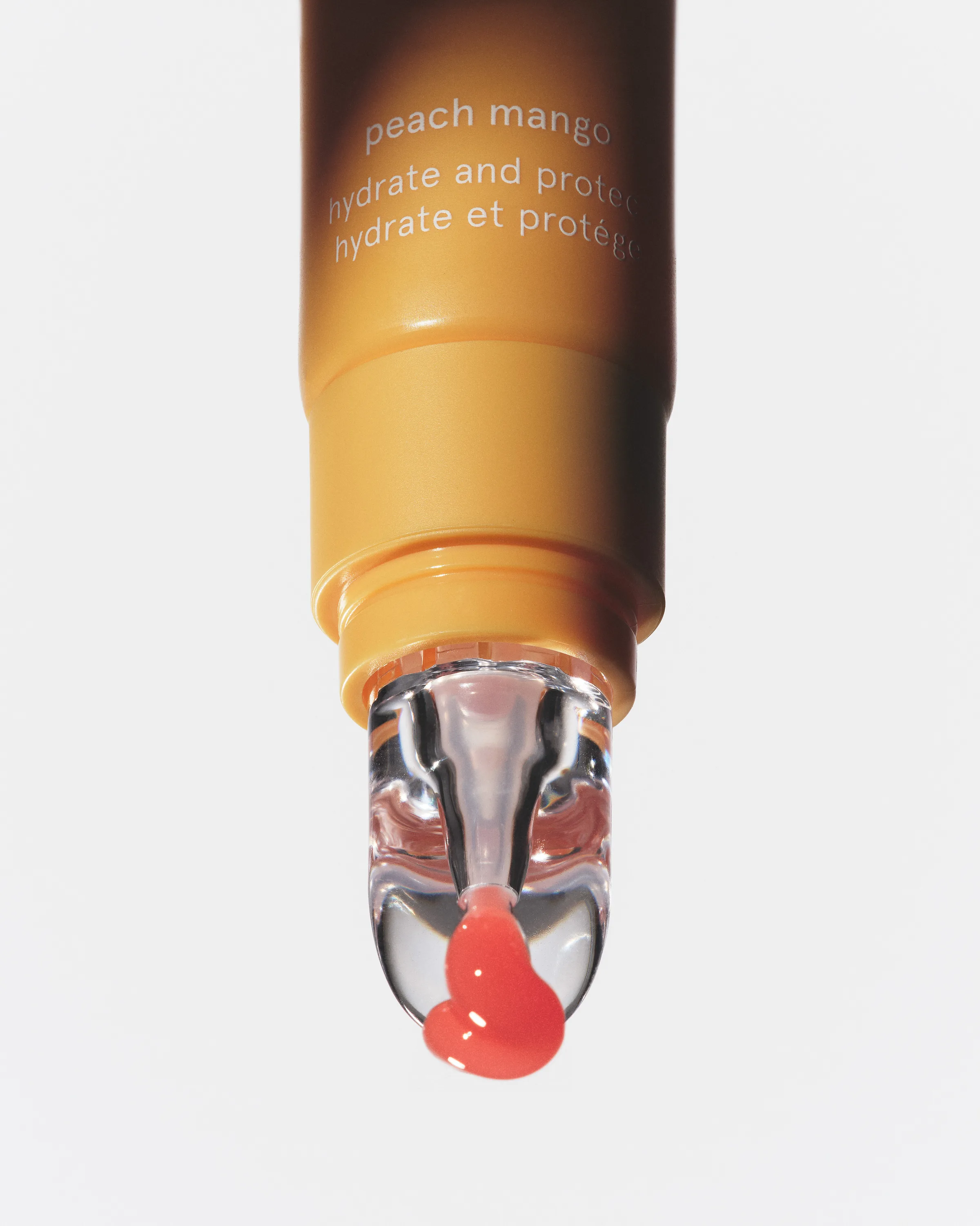 Close-up of a peach mango lip oil tube with a clear applicator dripping coral gloss on white, for Kylie Cosmetics.
