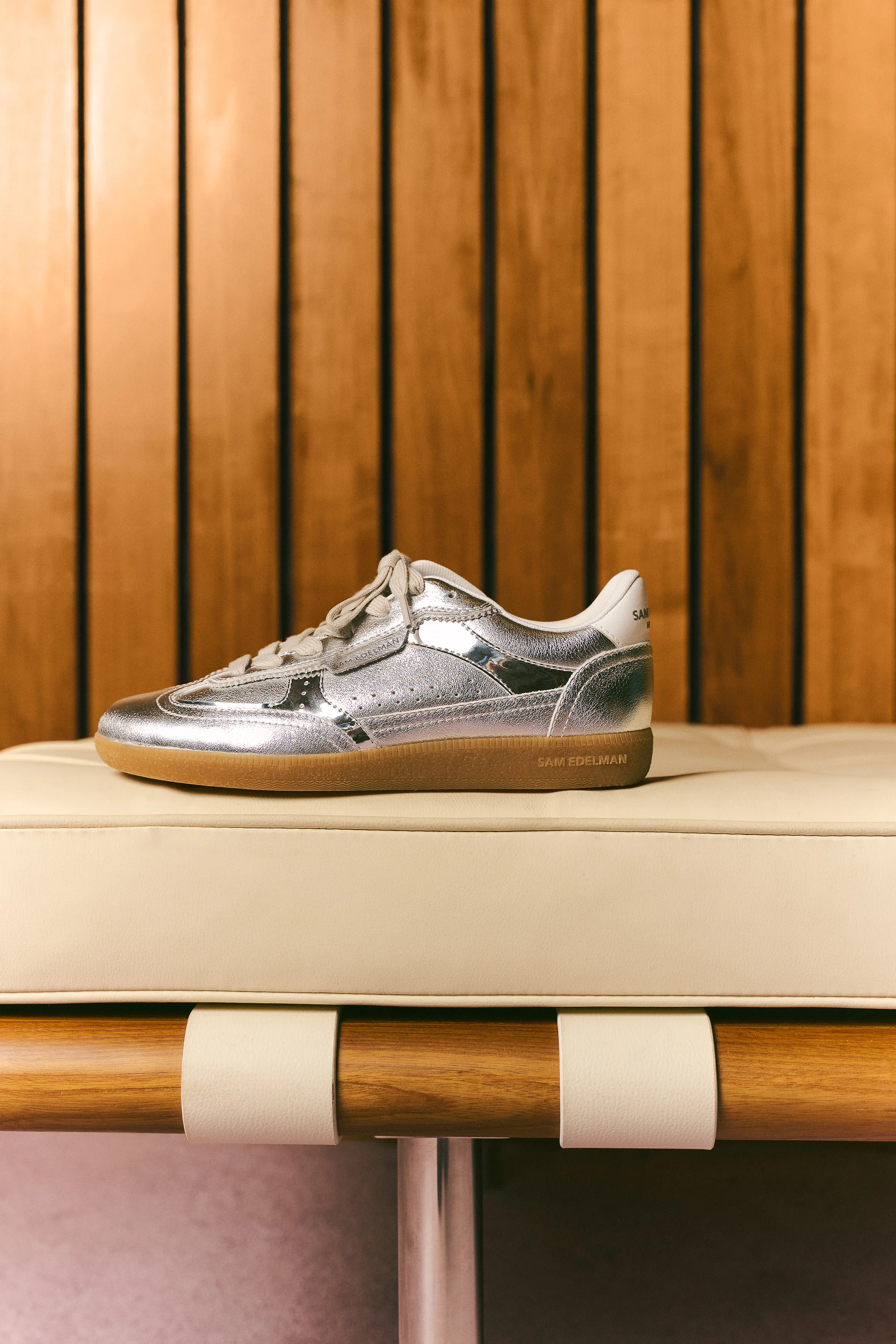 Silver metallic leather sneaker with gum sole on a cream cushioned bench before wood slat paneling, for Sam Edelman.