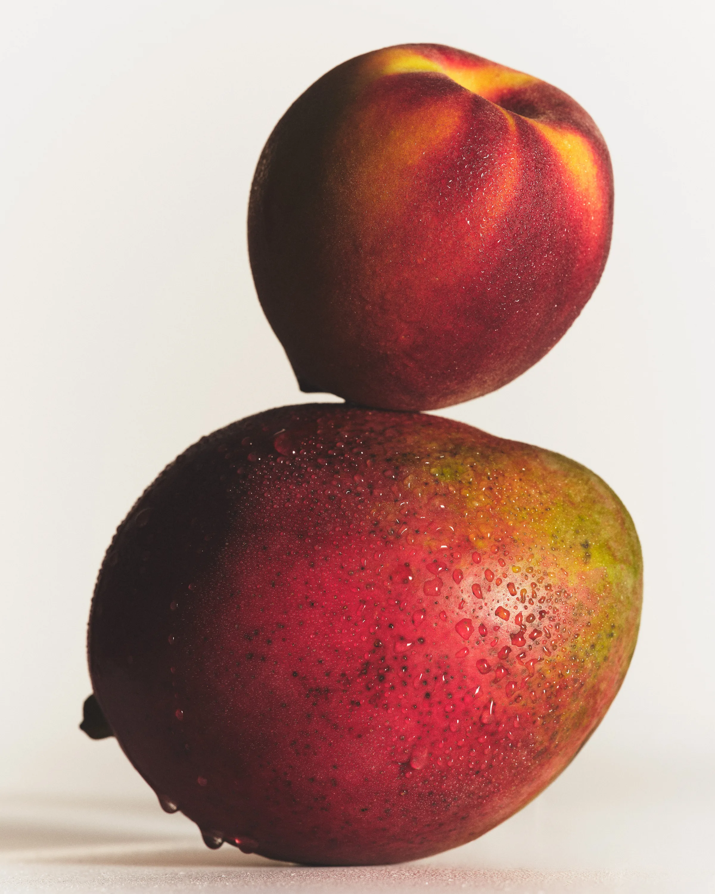 Still life of a ripe peach balanced on a water-beaded red mango against a pale backdrop, for Kylie Cosmetics.