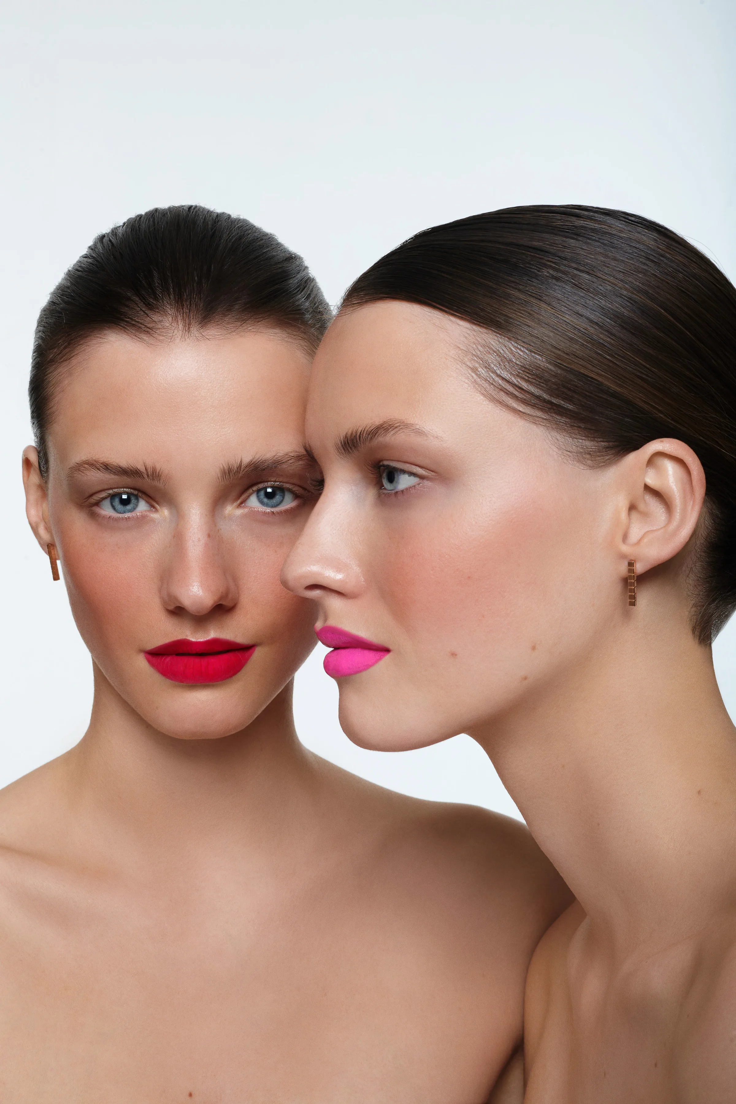 Two models with slicked-back hair, one wearing red matte lipstick, the other pink, cheek to cheek on a white background.
