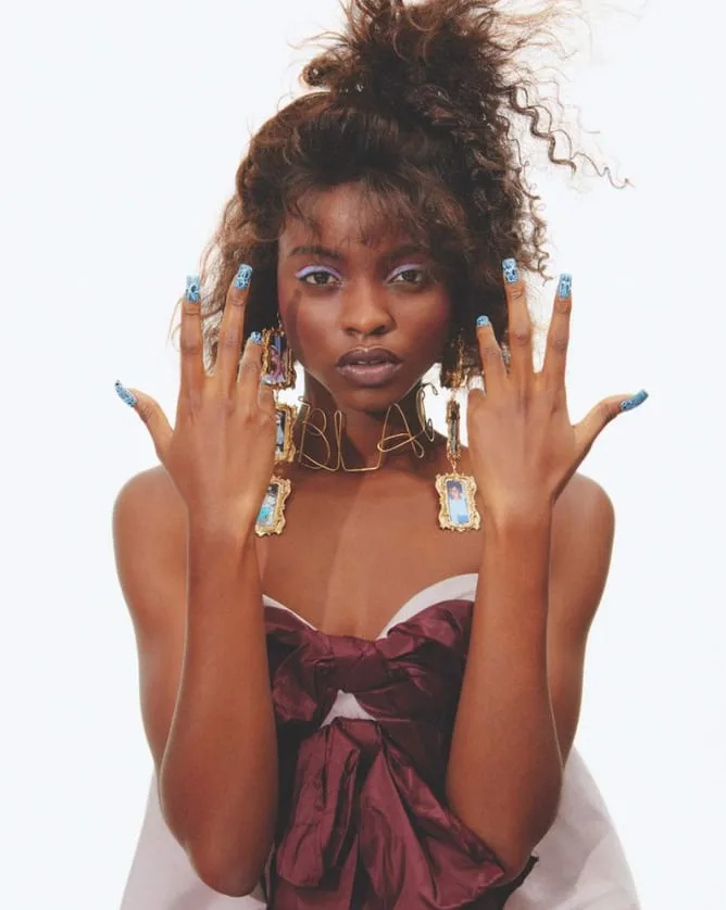 Model with curly updo shows blue patterned nails, gold frame earrings and wire necklace, in a burgundy bow top, for Office Magazine.