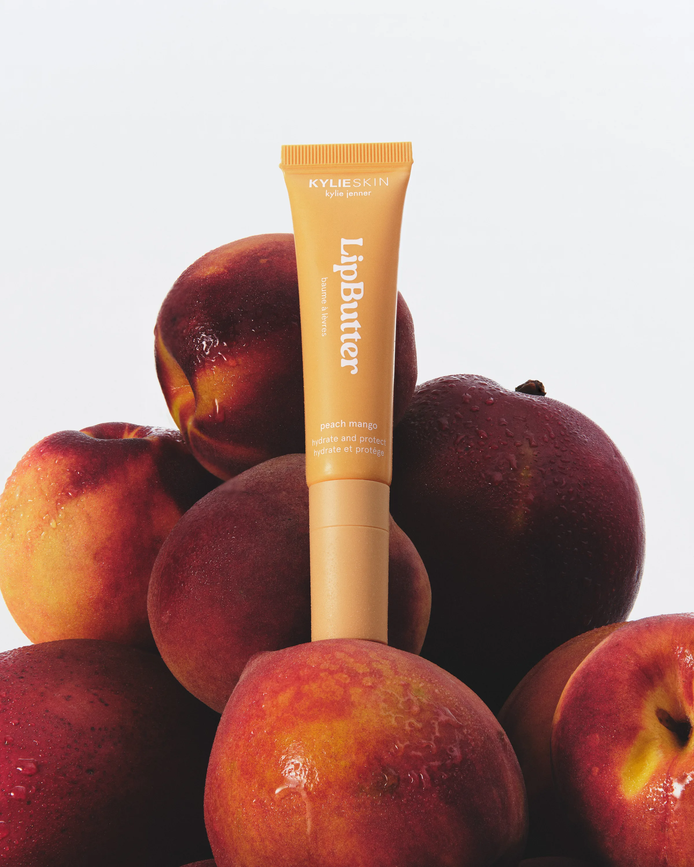 Peach mango Lip Butter tube standing among a pile of wet ripe peaches on a white background, for Kylie Cosmetics.