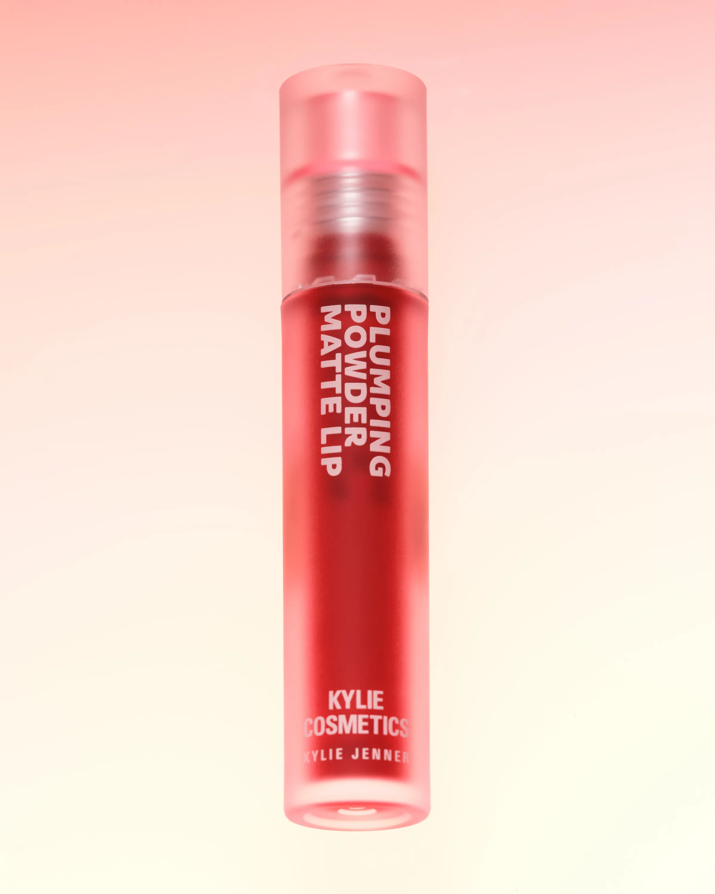 Red Plumping Powder Matte Lip tube with frosted pink cap on a soft pink-to-cream gradient, for Kylie Cosmetics.