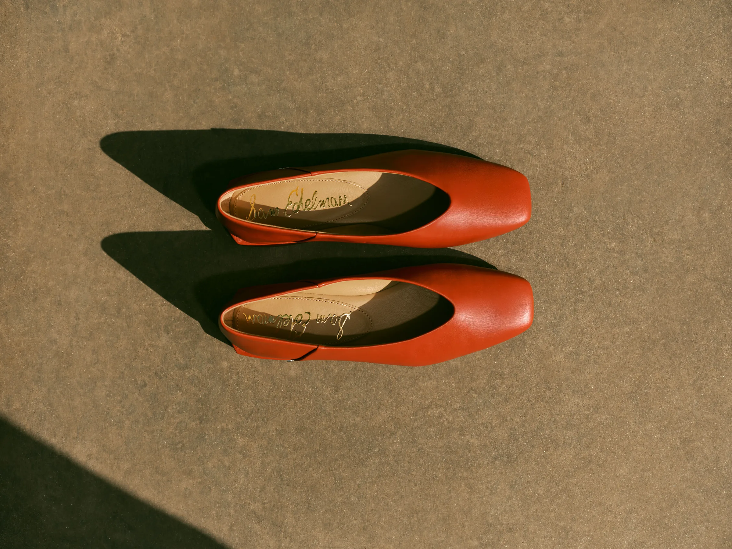 Pair of red leather square-toe flats casting long shadows on a sunlit concrete floor, shot from above, for Sam Edelman.