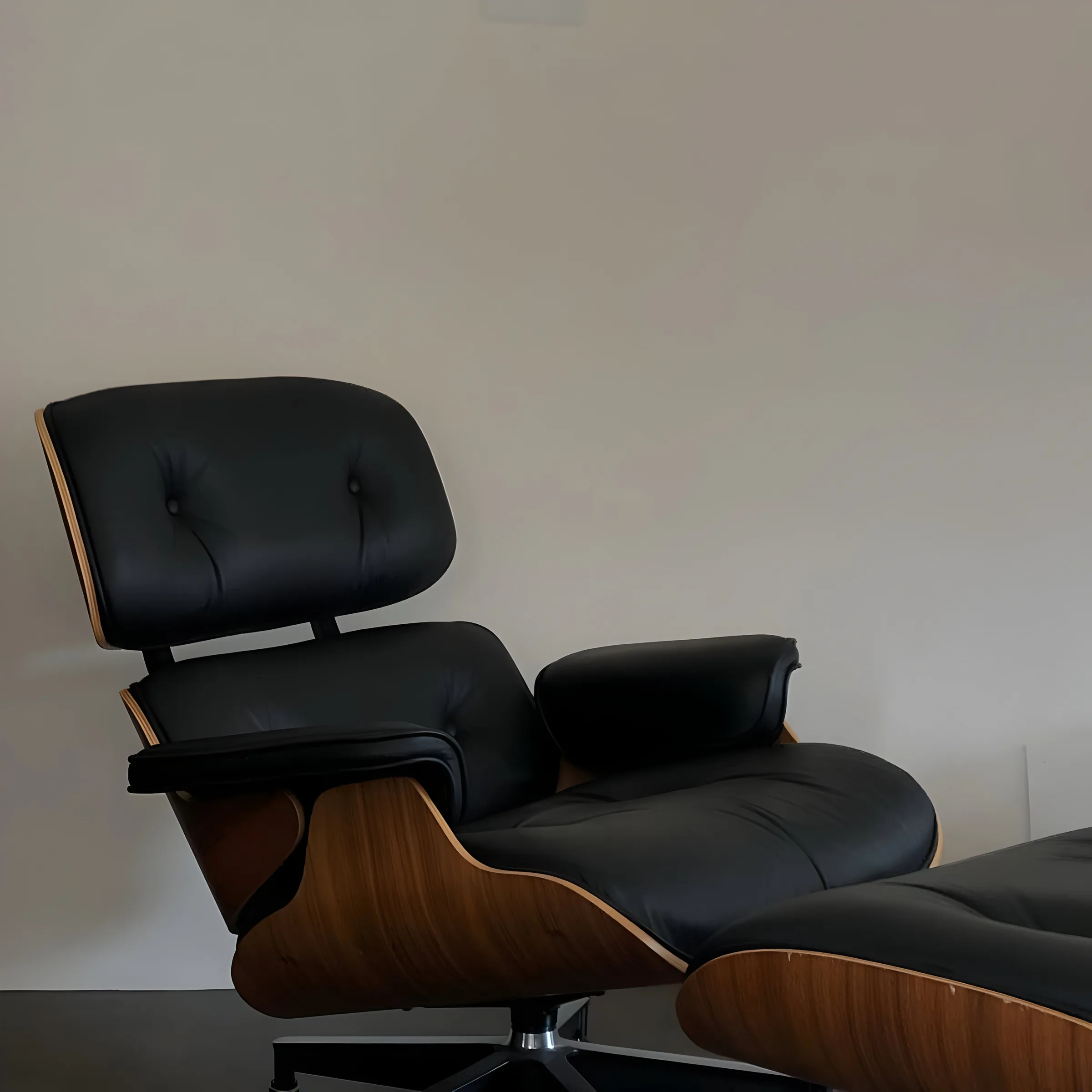 Black leather lounge chair with walnut shell and matching ottoman against a plain white wall.