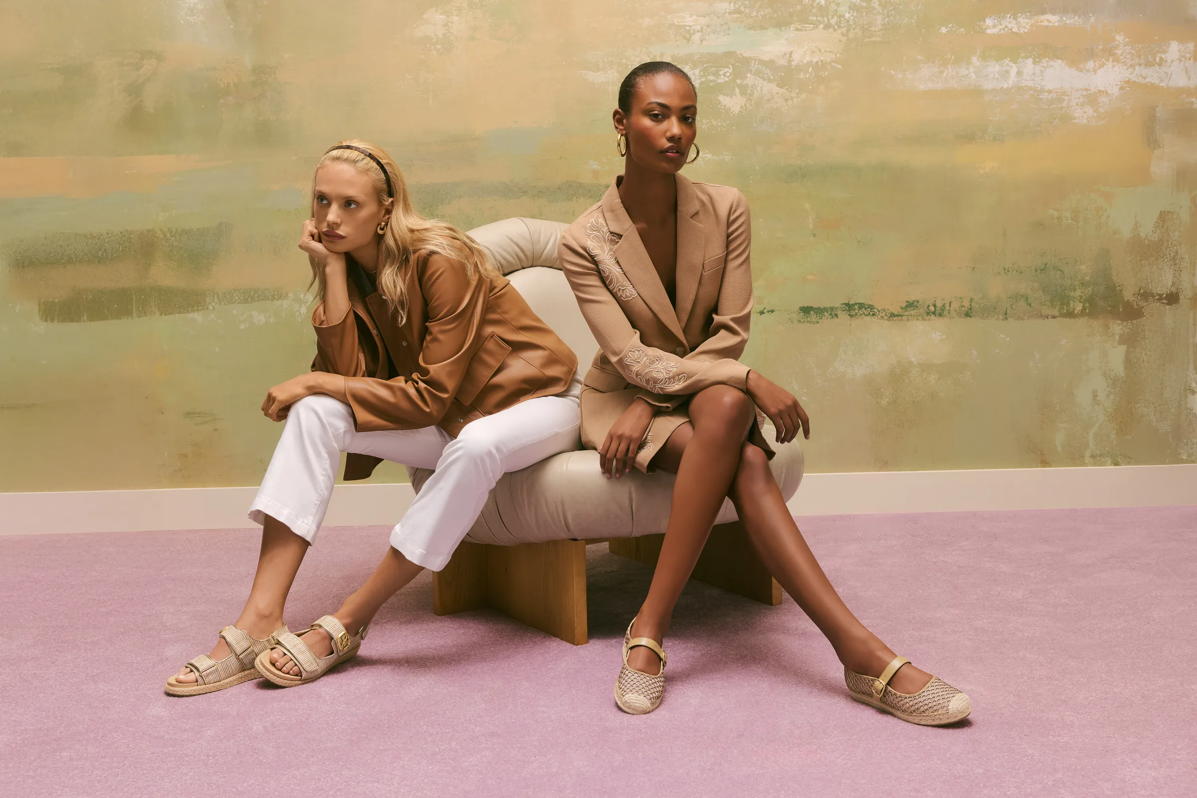 Two models in tan blazers pose on a cream chair against a painted wall, wearing mesh flats and sandals, for Sam Edelman.